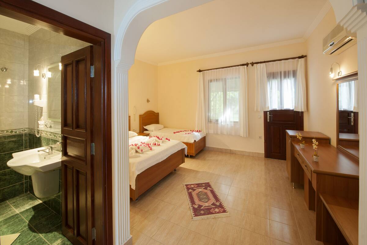 Habitación de hotel con baño, dos camas, un escritorio y un ventanal con cortinas.