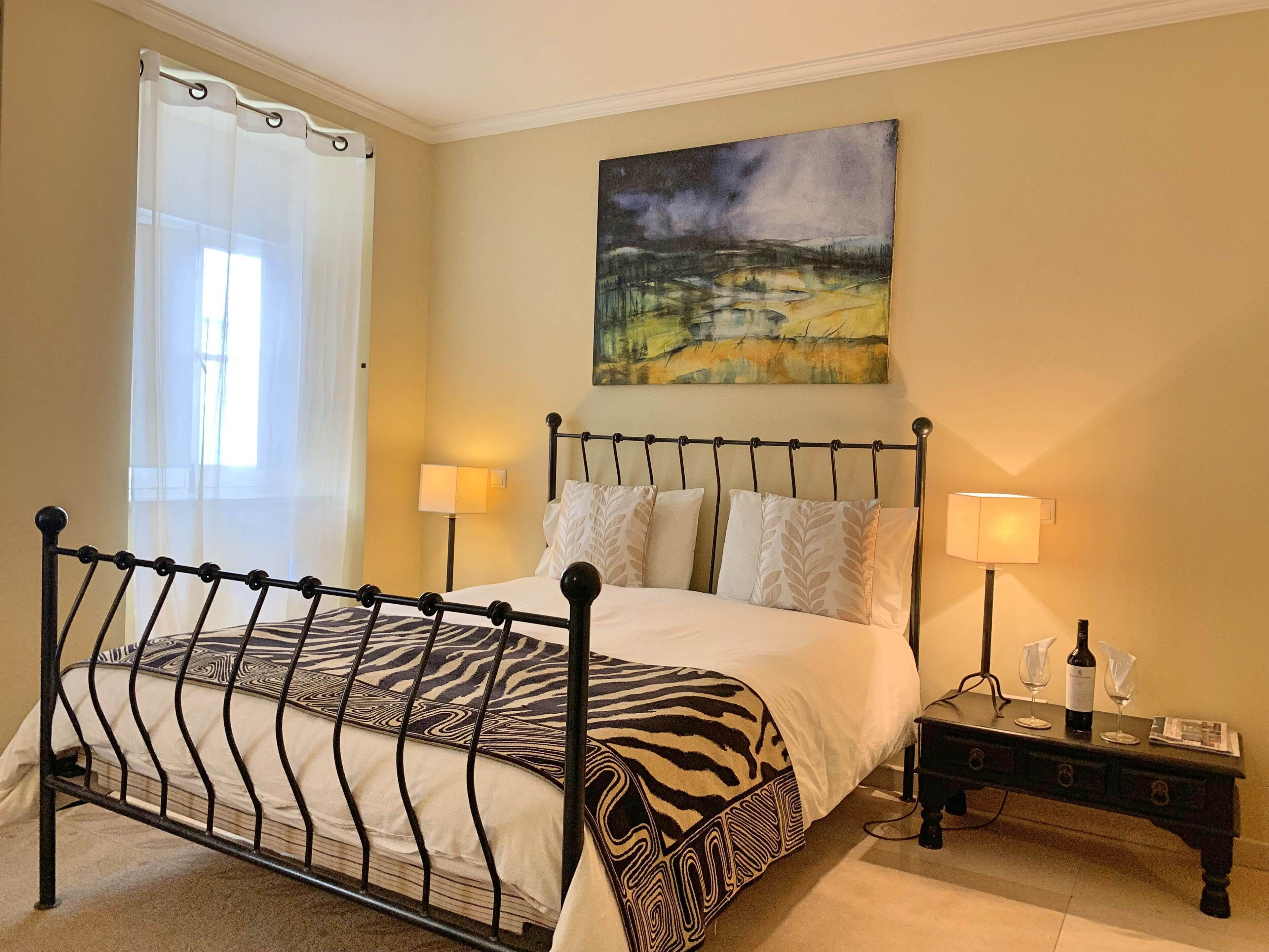 A bedroom with a zebra-patterned bedspread, two bedside lamps, a painting on the wall, and a nightstand with a bottle and glasses.