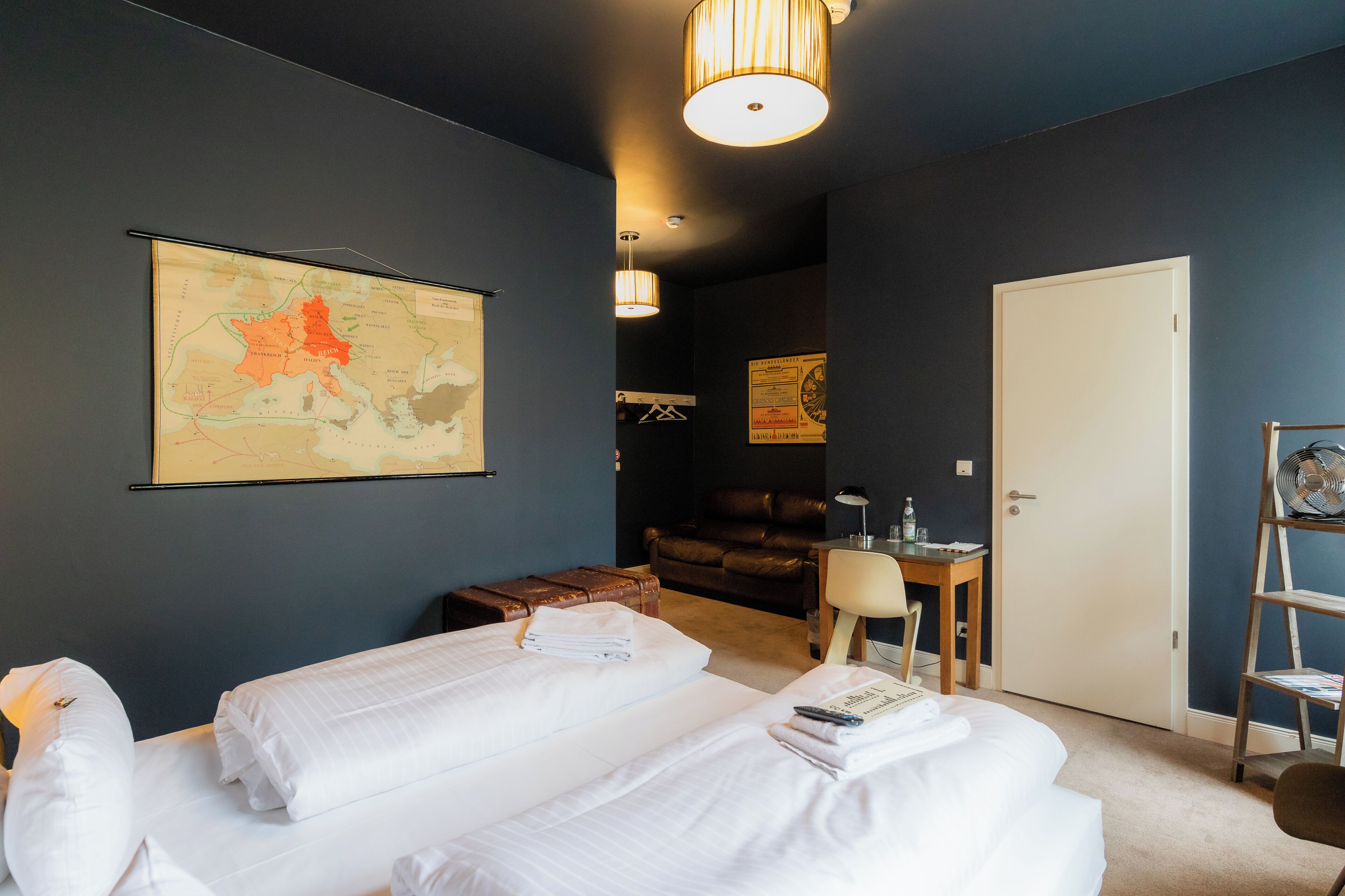 A hotel room with two beds, a map on the wall, a desk with a chair, and a door.