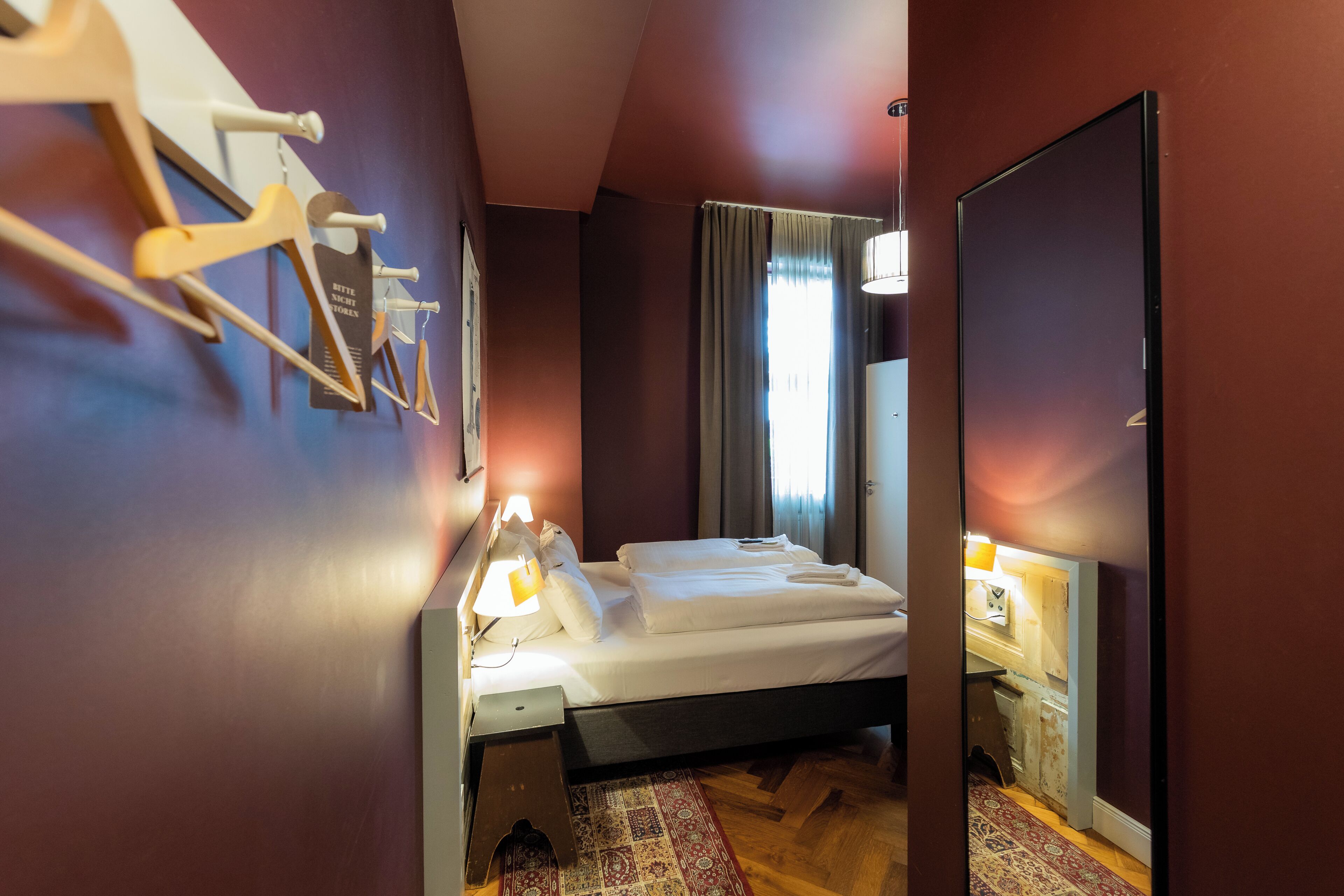 Una habitación de hotel con cama, escritorio con lámpara, espejo y ventana con cortinas.