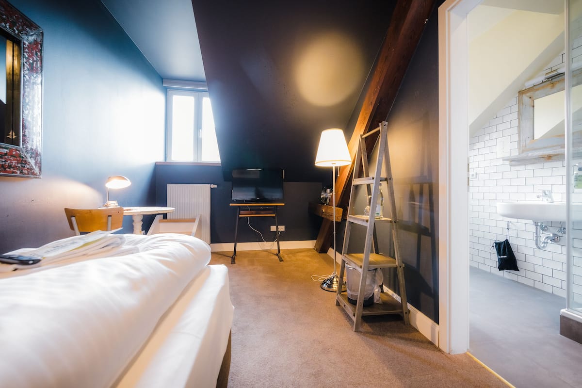 A hotel room with a bed, a desk, a lamp, a ladder, and a bathroom with a sink and mirror.