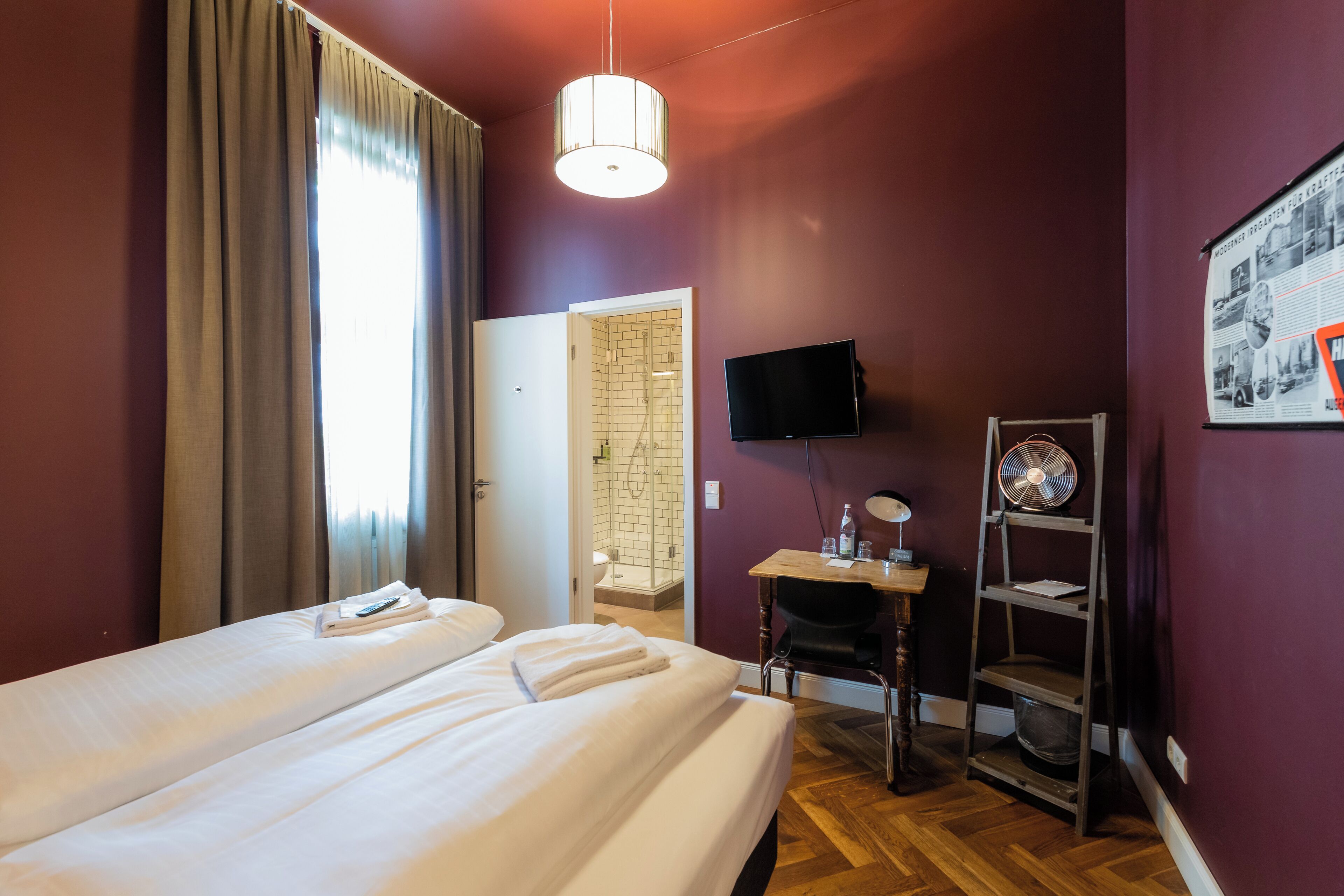 A hotel room with two beds, a desk with a chair, a TV, and a fan.