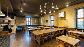 A restaurant interior with wooden tables, hanging pendant lights, a clock on the wall, and a view through the window.