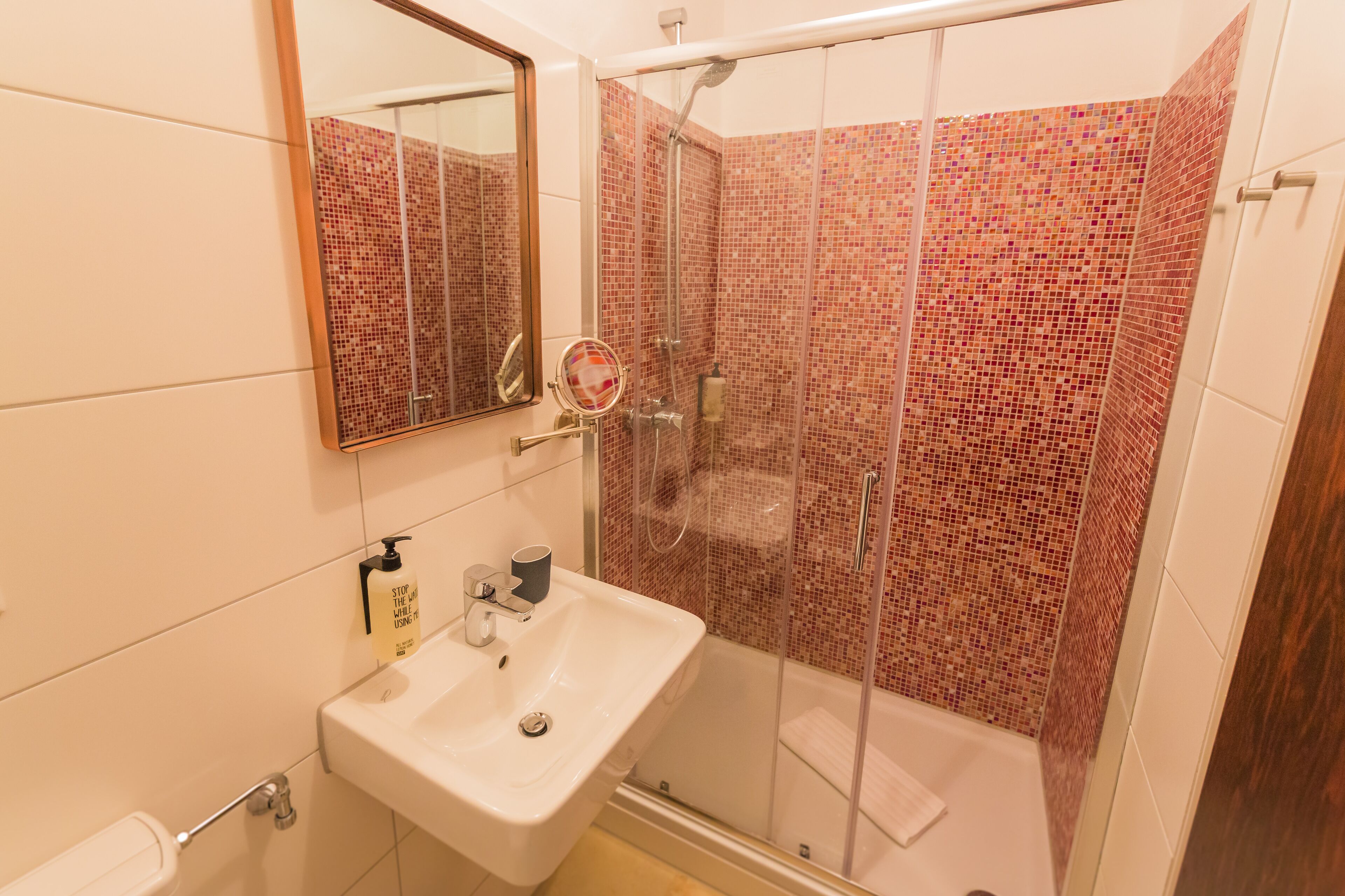 Un baño con ducha envuelta en cristal, un lavabo con espejo y pared de azulejos.