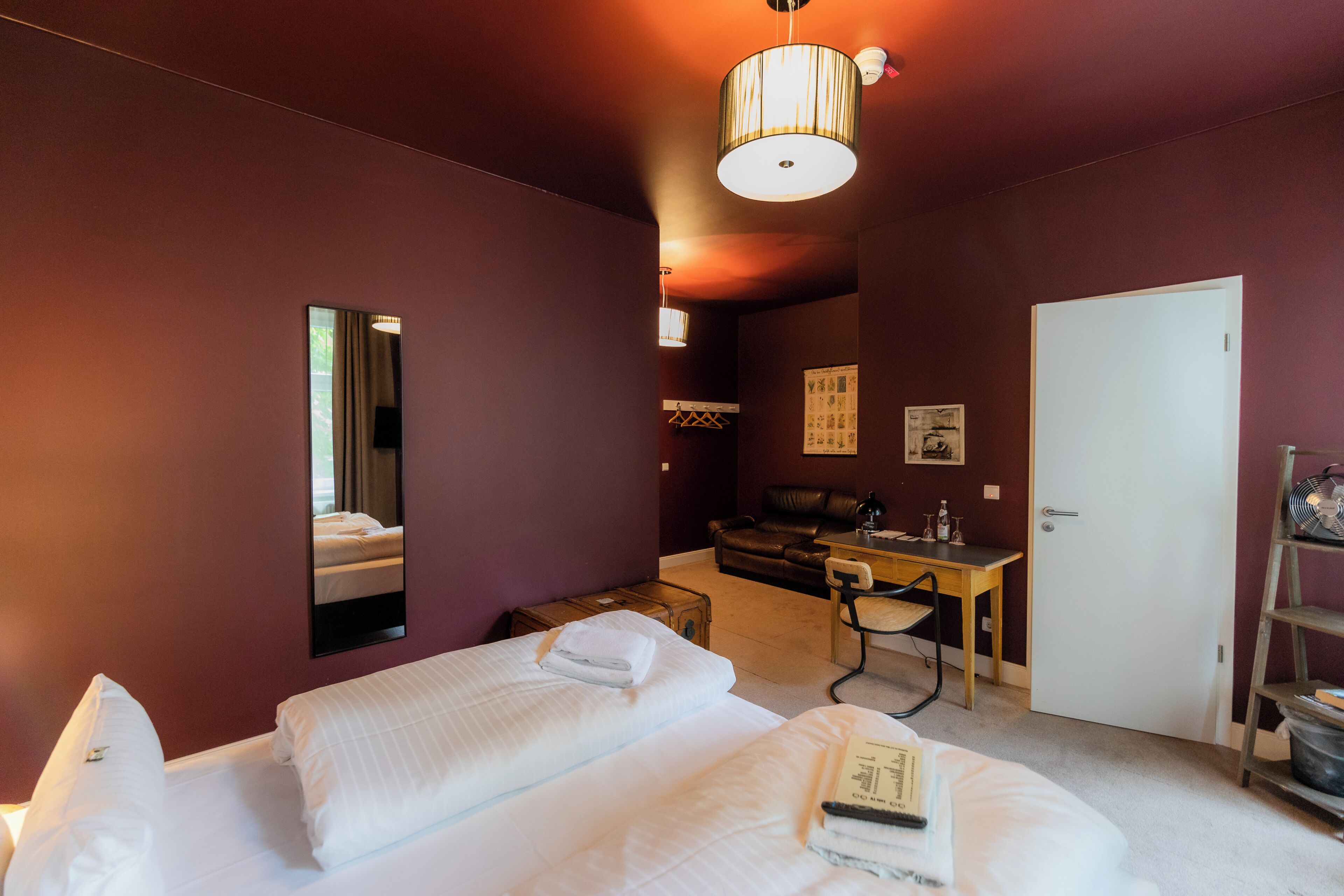 Habitación de hotel con cama, escritorio, silla, espejo, puerta y una pequeña mesa con una botella.