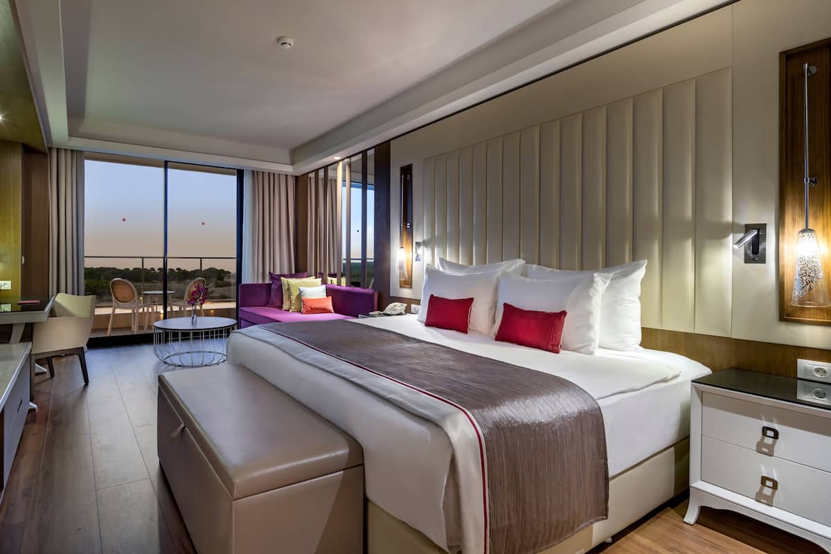 A modern hotel room with a large bed, a desk, a sofa, and a balcony with a view.