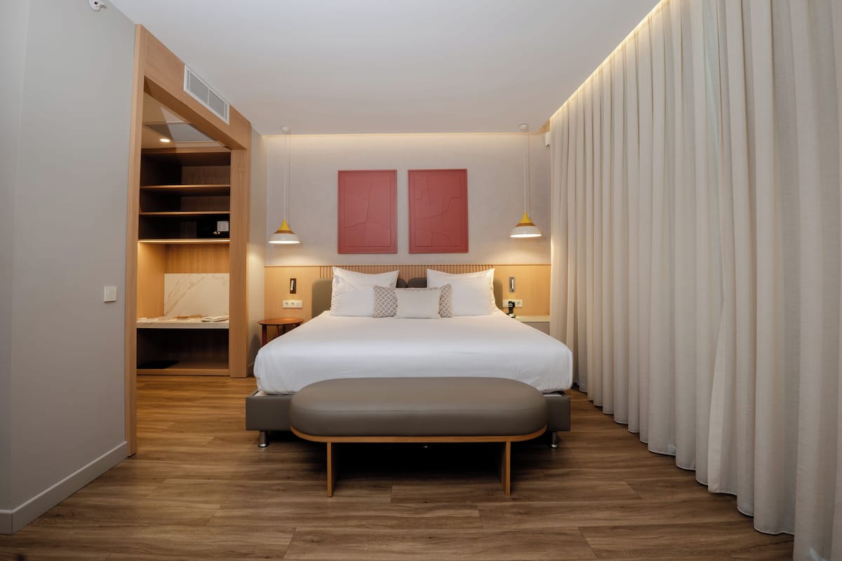 A modern hotel room with a large bed, wooden flooring, a bench, a wardrobe, and two wall-mounted lamps.