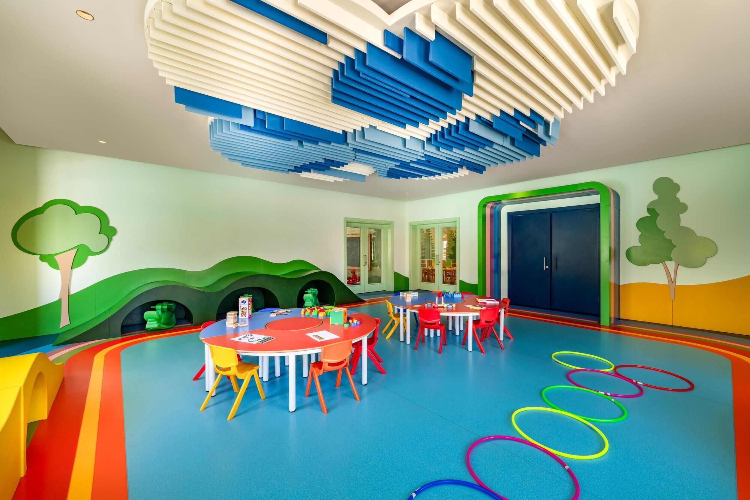 A brightly colored children's playroom with tables and chairs, a colorful floor design, and playful wall decorations.