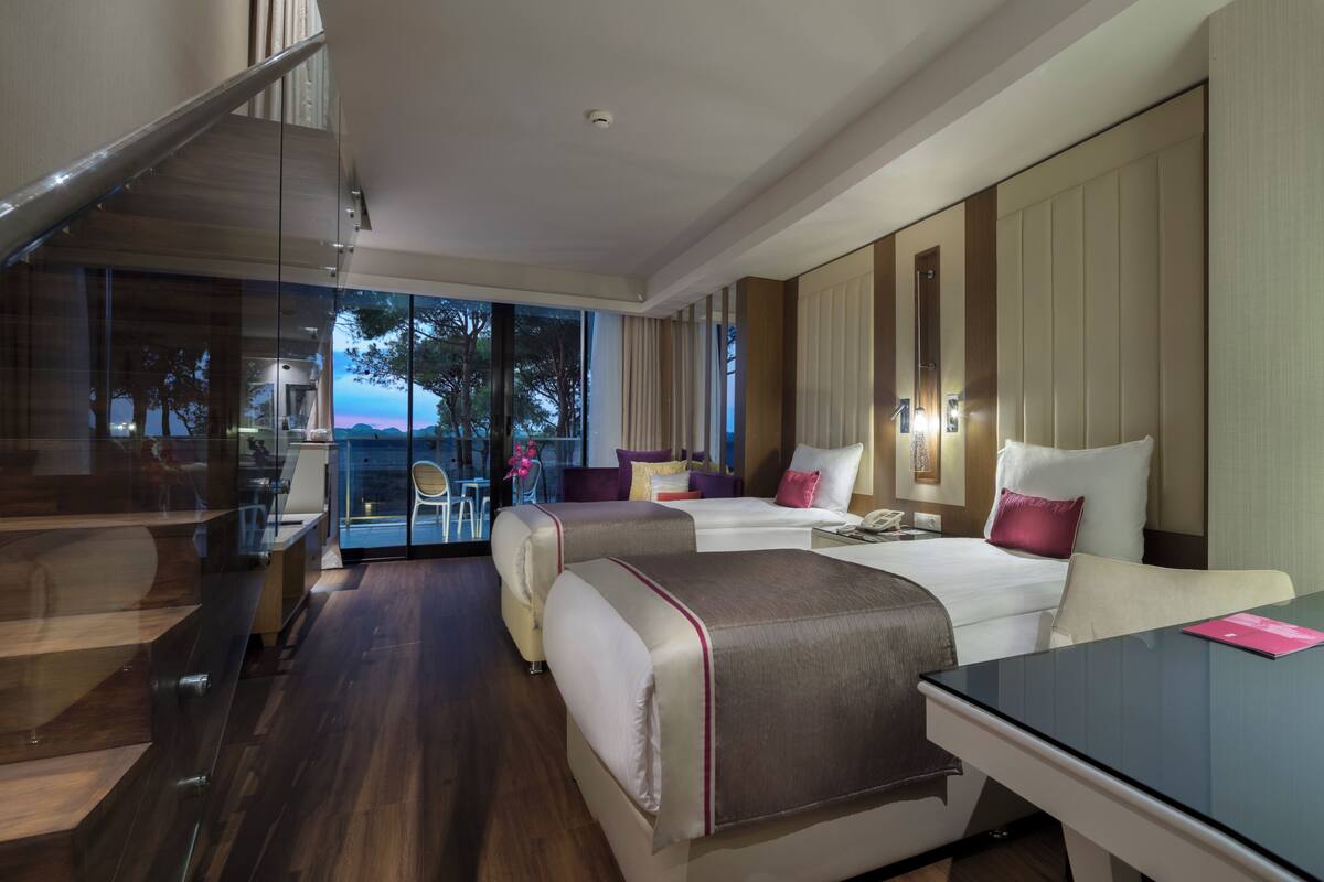 A modern hotel room with a glass staircase, a balcony with a view, two beds, a desk, and a chair.