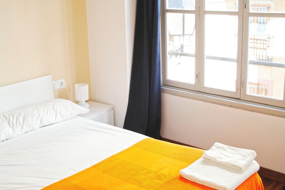 A hotel room with a bed, a nightstand, a window with curtains, and a view of buildings outside.