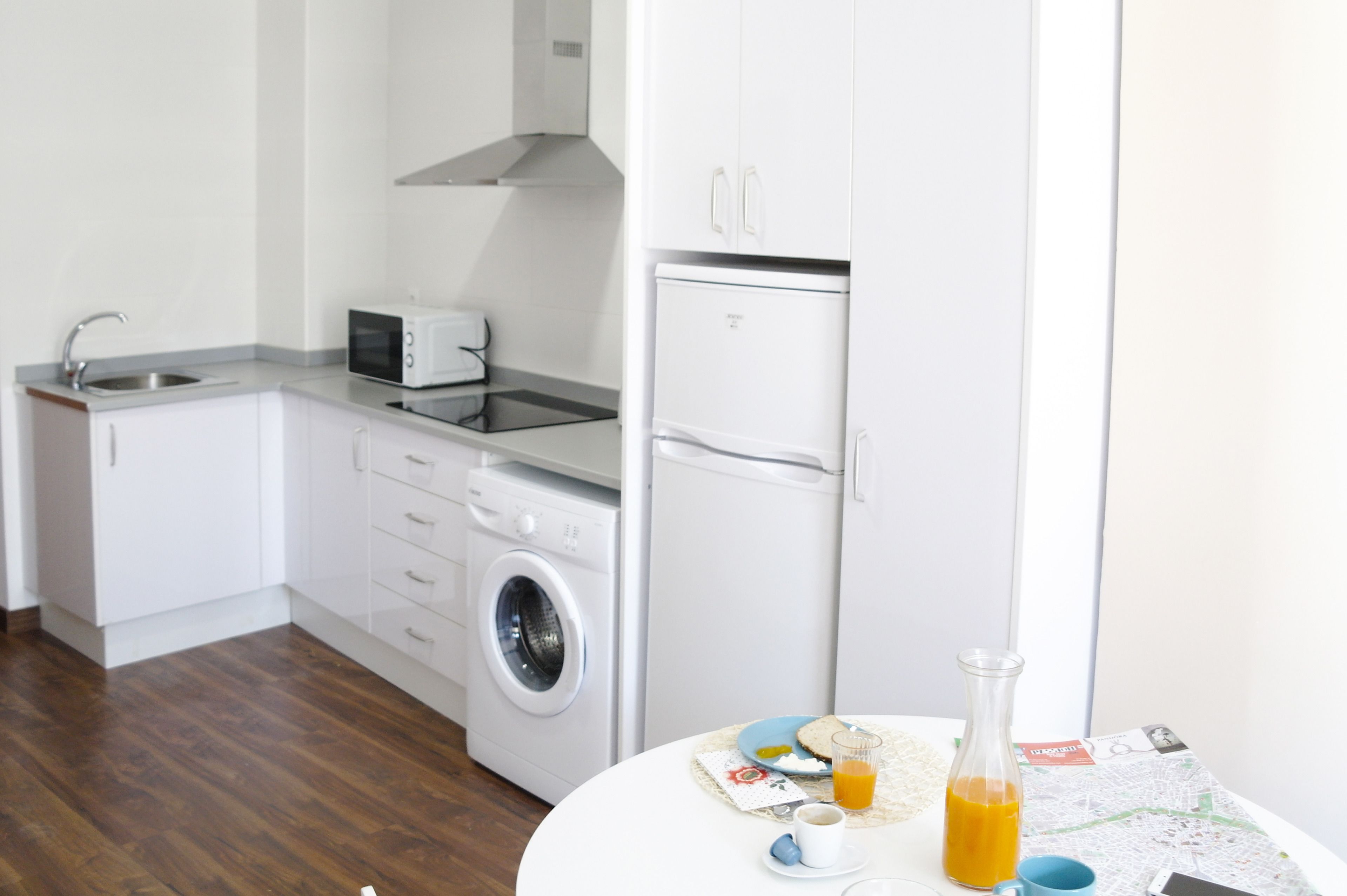 A compact kitchen with a washing machine, a microwave, and a refrigerator. There is a table with a bottle of orange juice, a cup, and a plate with food.