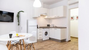 Une cuisine moderne avec des meubles blancs, un lave-linge et un four à micro-ondes. On y trouve une table à manger avec des chaises et un téléviseur fixé au mur.