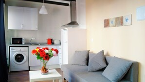 Une cuisine moderne avec des meubles blancs, un four à micro-ondes et un lave-linge. On y trouve un canapé gris avec des coussins et un vase de fleurs posé sur une petite table.