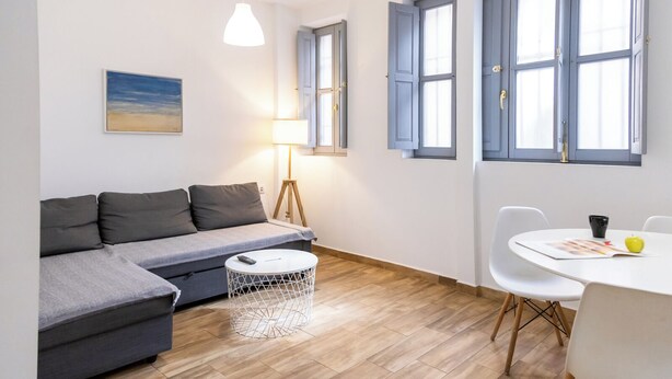 Un salon moderne avec un canapé gris, une table basse ronde et une table à manger avec des chaises. On y trouve une lampe, un tableau au mur et de grandes fenêtres avec des volets bleus.