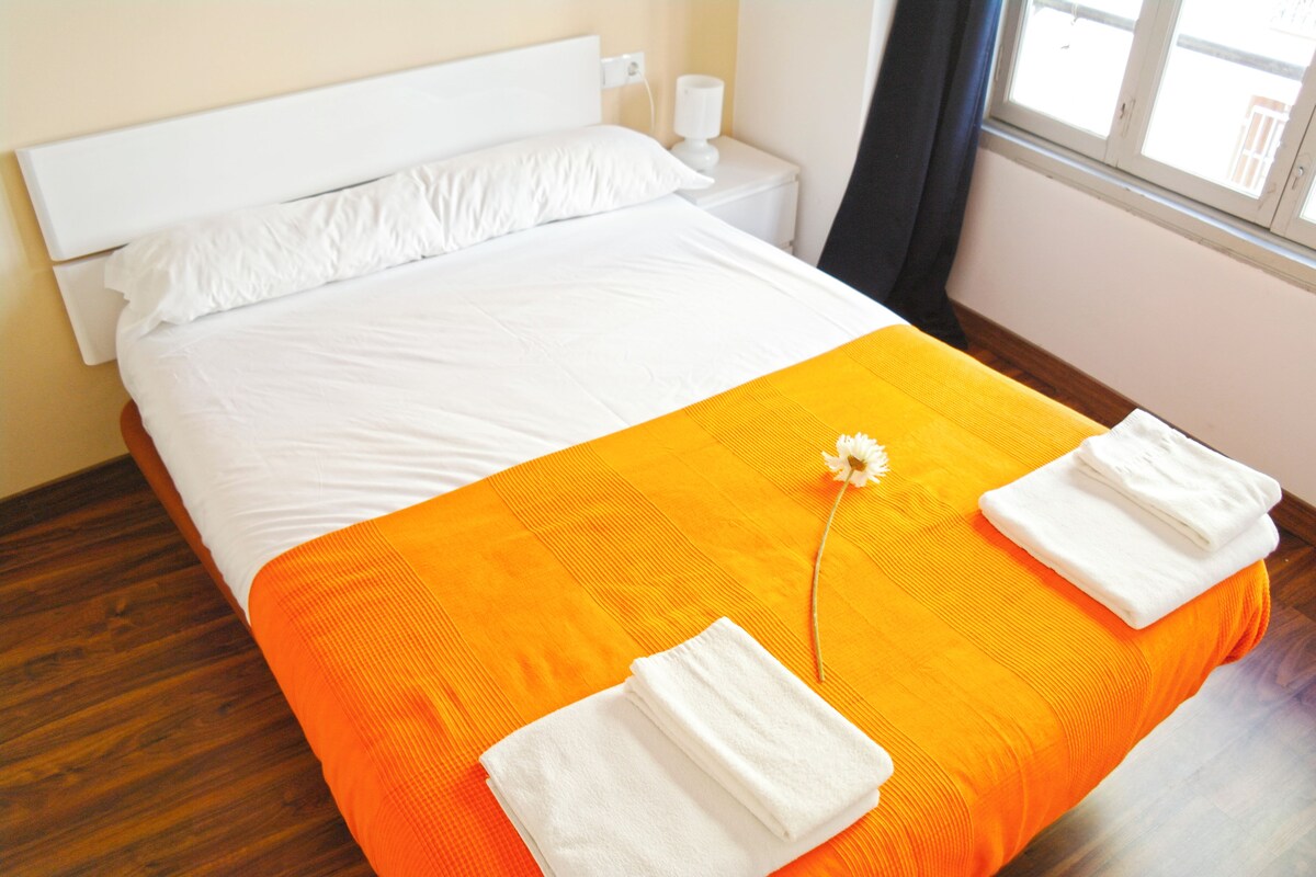 A bed with white linens and an orange bedspread, a white pillow, and a white towel on the side.