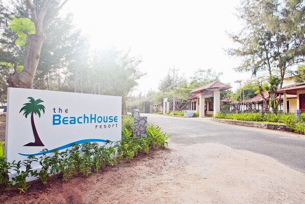 A sign for The BeachHouse Resort with a palm tree logo, surrounded by greenery and a paved road. - The Beach House Resort (Ho Chi Minh City)
