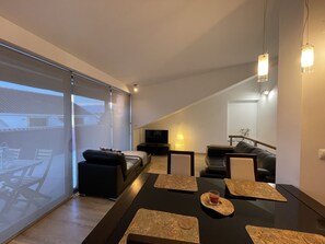 Ein modernes Wohnzimmer mit einer großen Schiebeglastür, einem schwarzen Ledersofa, einem Esstisch mit Untersetzern und einem Fernseher auf einem Lowboard.