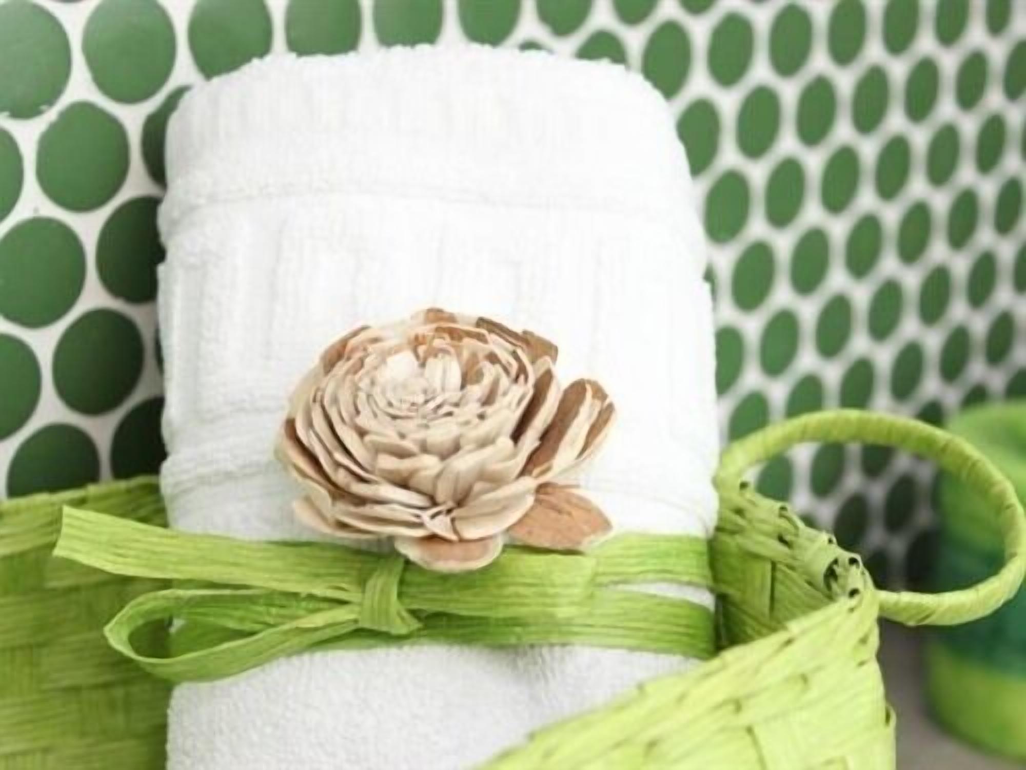 A white towel with a beige flower decoration, tied with a green ribbon, placed in a green woven basket.