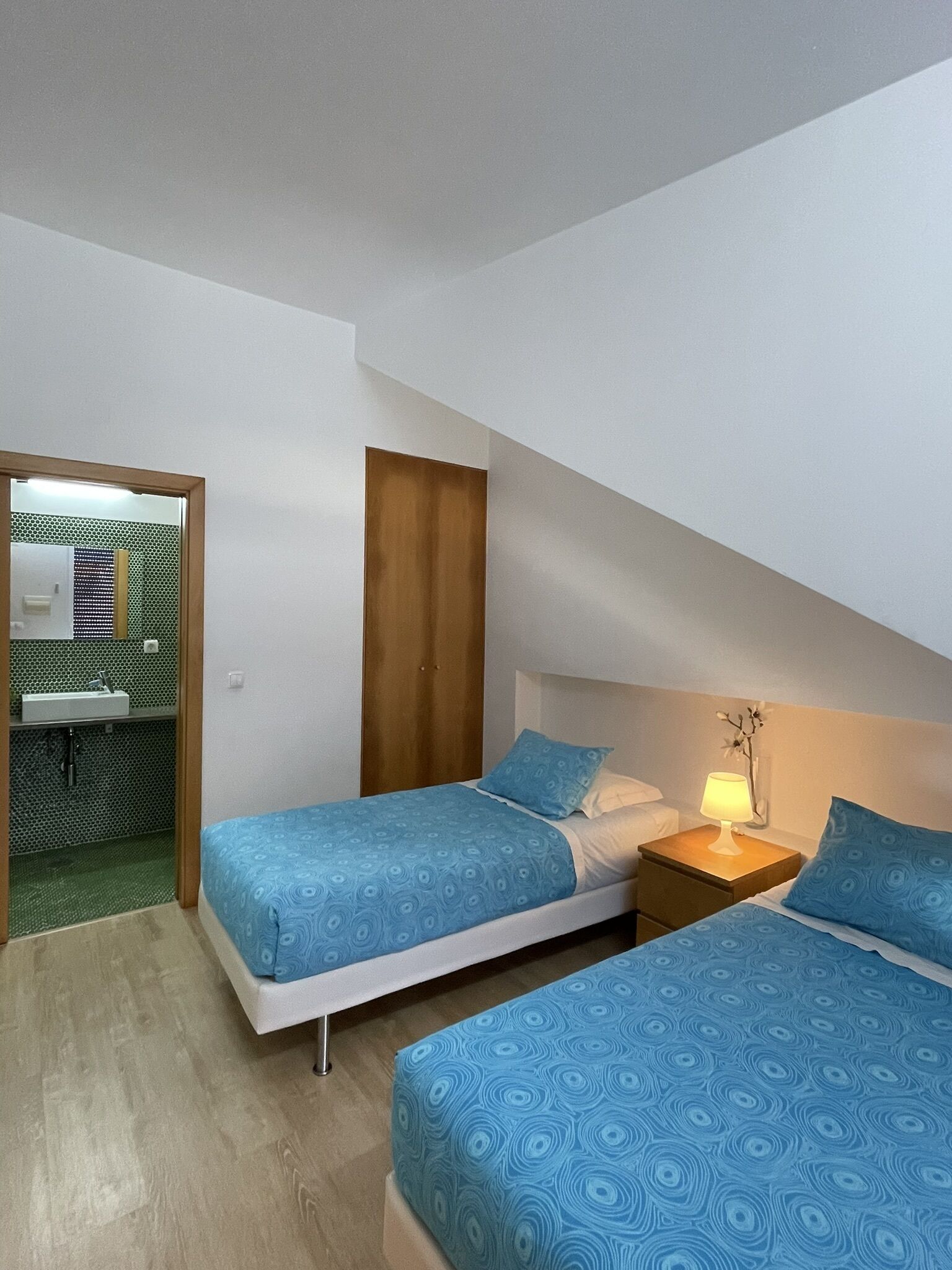Quarto com duas camas, uma mesa de cabeceira, uma luminária e banheiro visível por uma porta aberta.