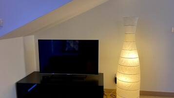 Een kamer met een flatscreen televisie op een zwarte standaard, een hoge, cilindrische lamp en een houten vloer.