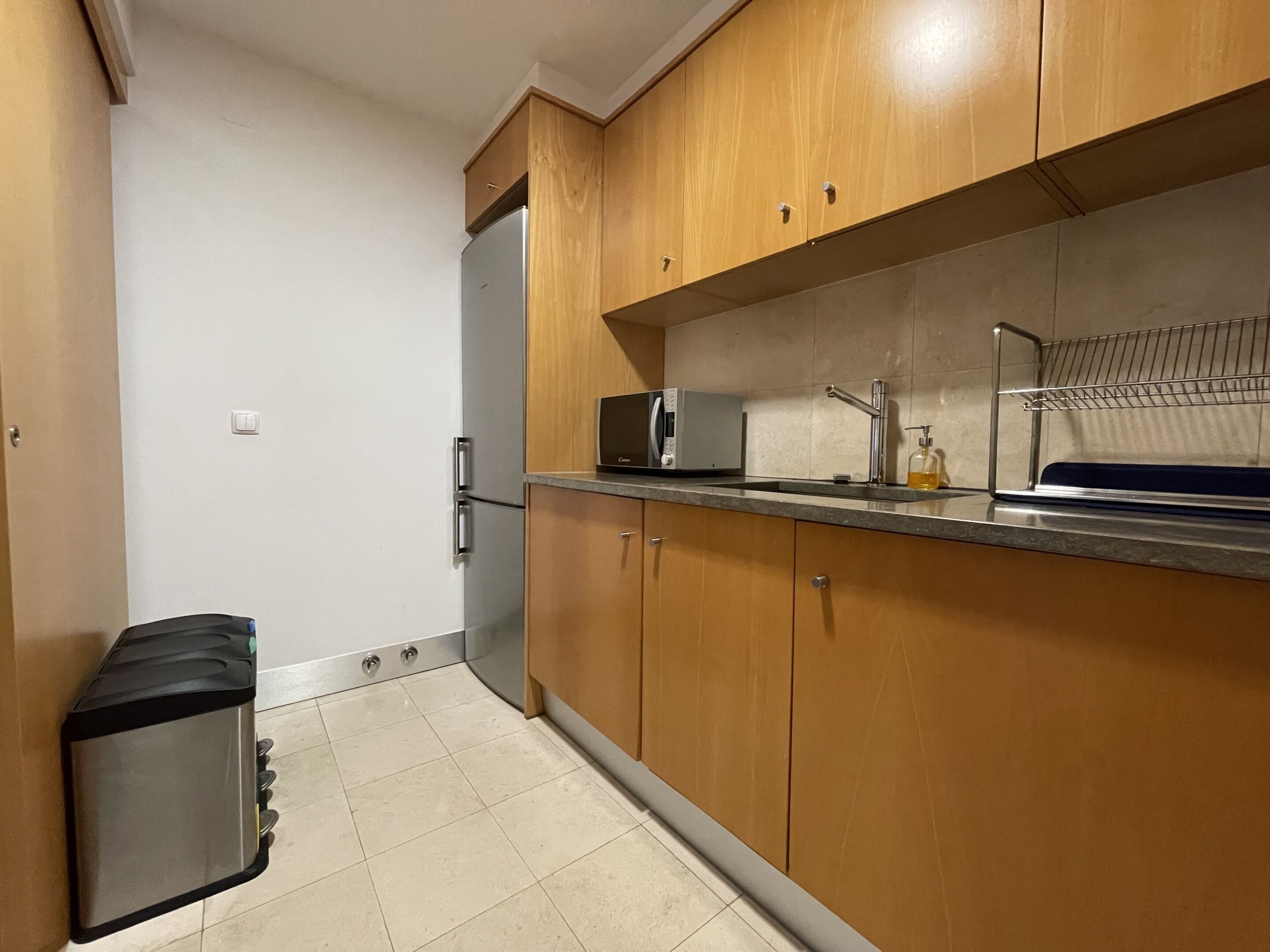 A compact kitchen with wooden cabinets, a stainless steel refrigerator, a microwave, and a sink with a soap dispenser.