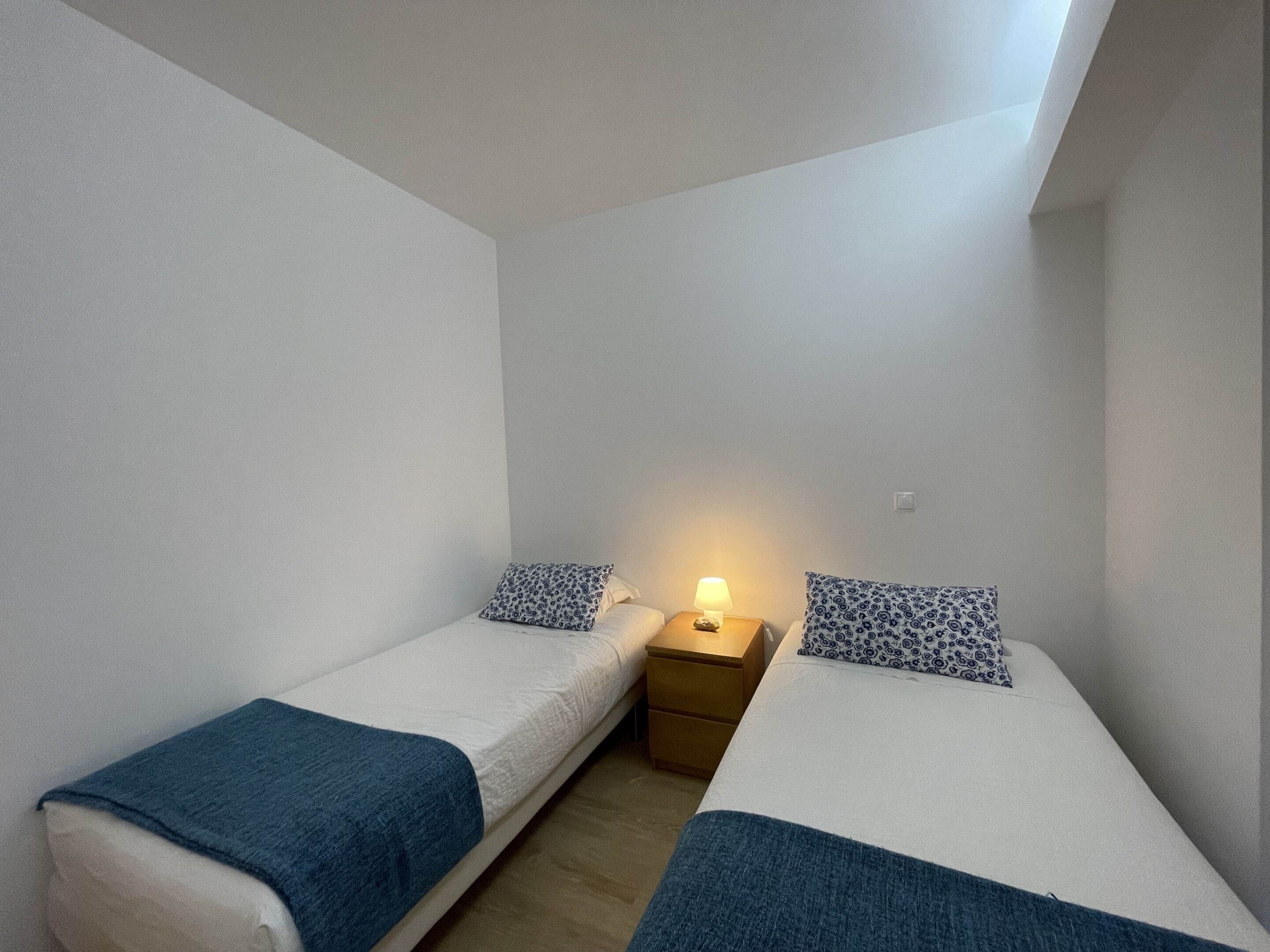 Il s’agit d’un petit logement composé d’une seule pièce, avec deux lits simples, chacun agrémenté d’un coussin à motifs et d’une couverture bleue. Une table de chevet avec une lampe se trouve entre les deux lits.