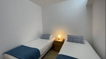 Il s’agit d’un petit logement composé d’une seule pièce, avec deux lits simples, chacun agrémenté d’un coussin à motifs et d’une couverture bleue. Une table de chevet avec une lampe se trouve entre les deux lits.