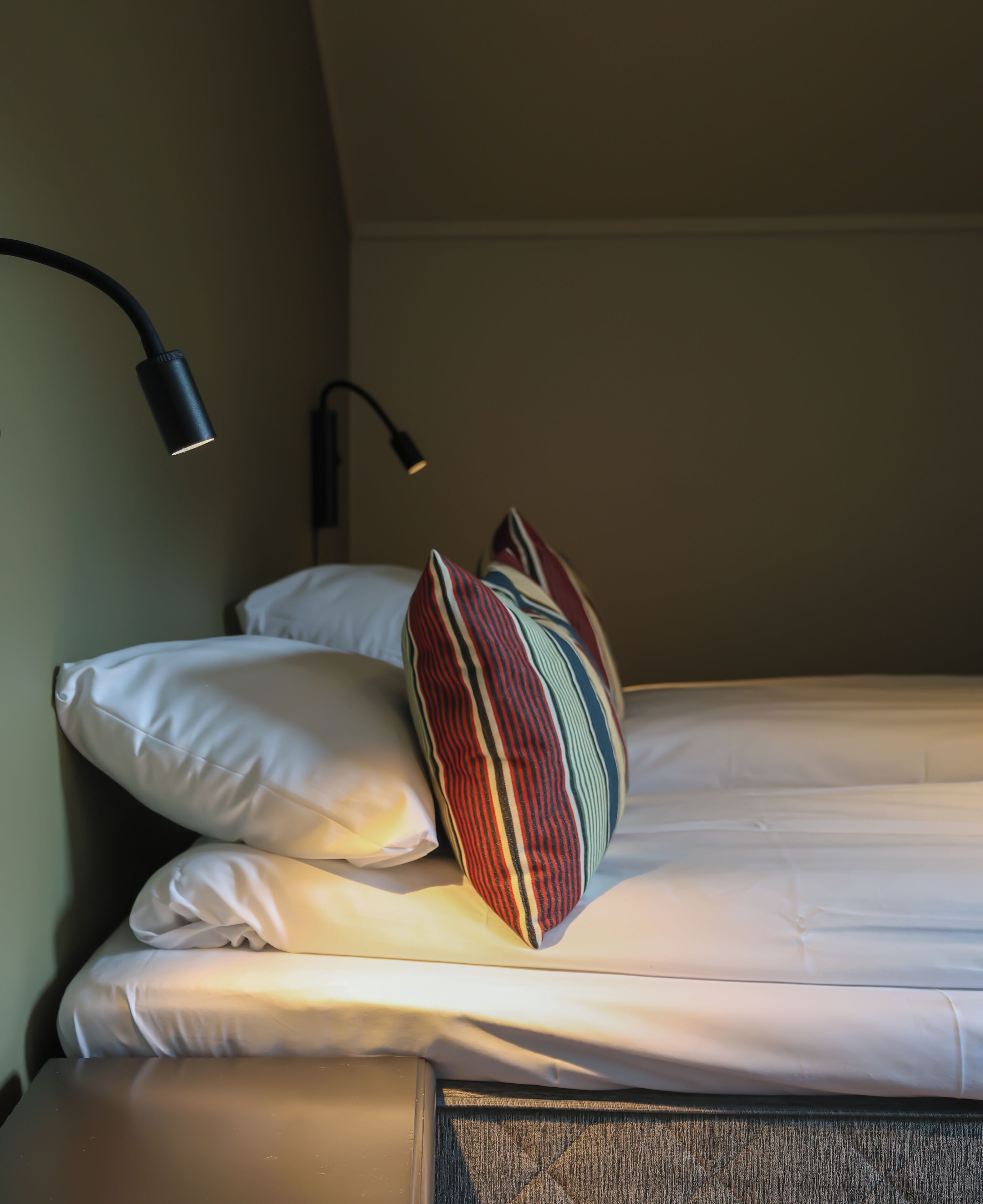 Un lit bien fait, avec du linge de lit blanc, un coussin à rayures et une lampe noire fixée au mur.