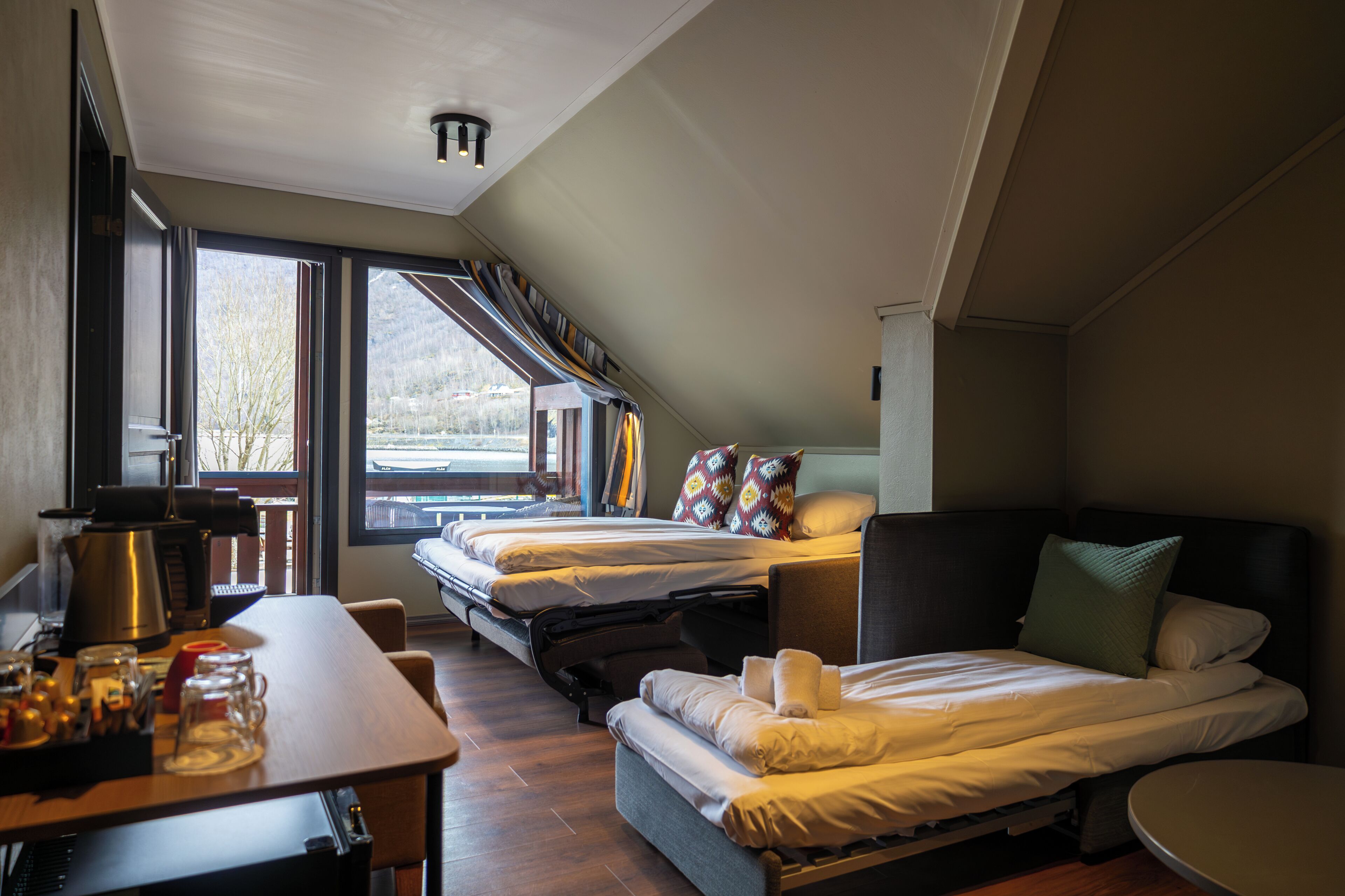 Una habitación de hotel moderna con dos camas, un área de comedor con mesa y sillas, y ventanas grandes que ofrecen vistas al exterior.