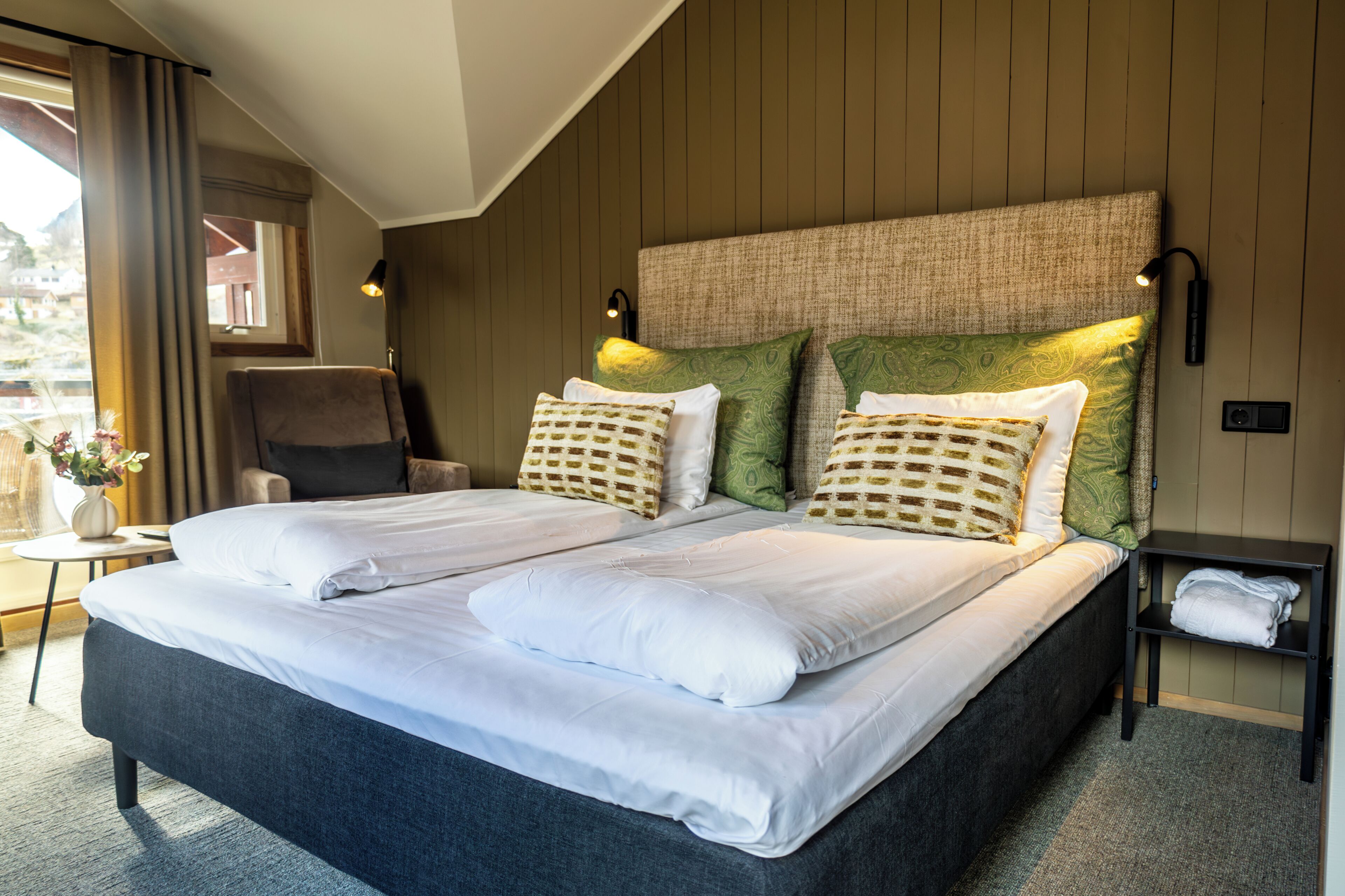 A double bed with white linens, a headboard with a textured pattern, two wall-mounted reading lamps, and a small bedside table with a folded towel.