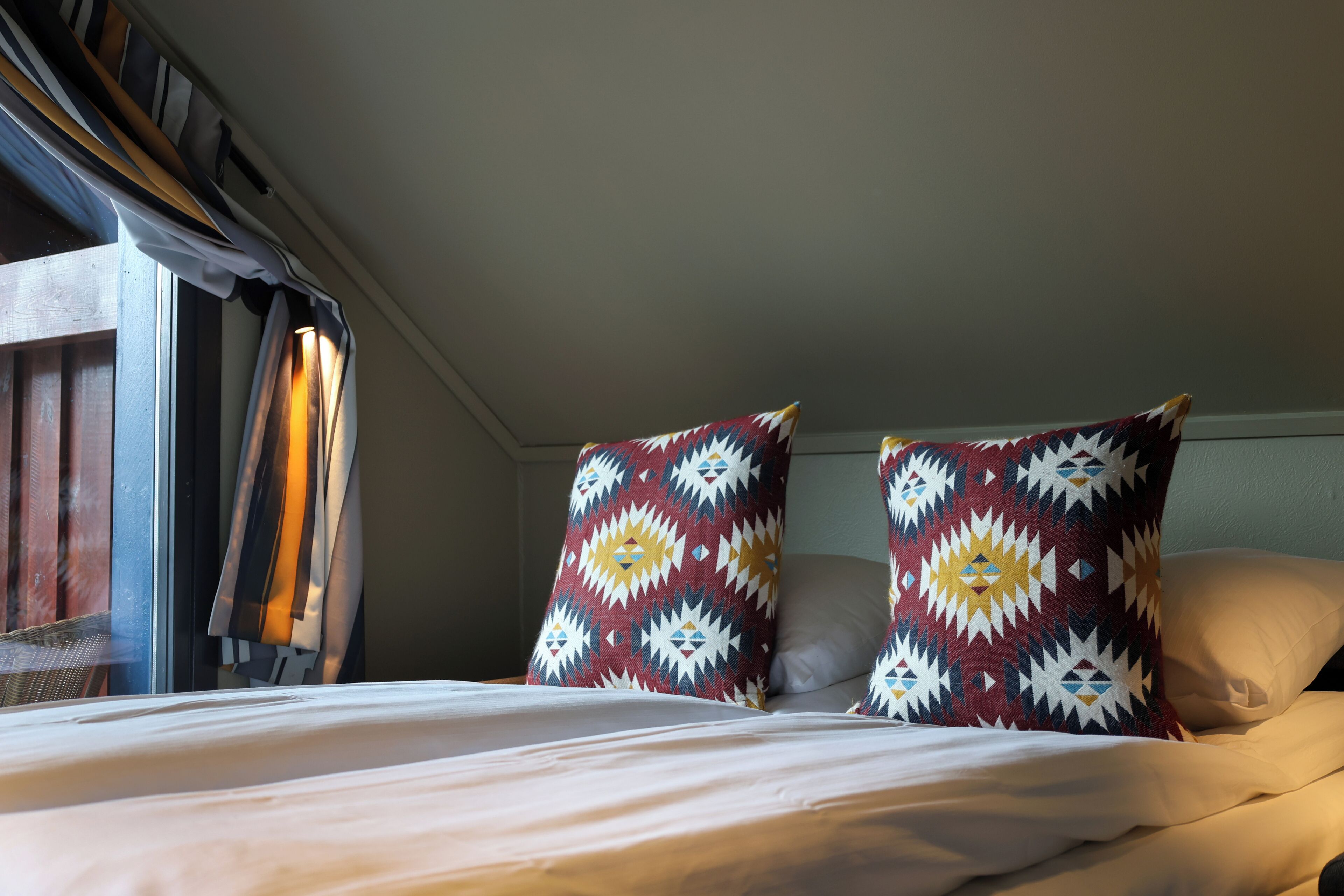 A bed with white bedding, two patterned pillows, and a window with curtains.
