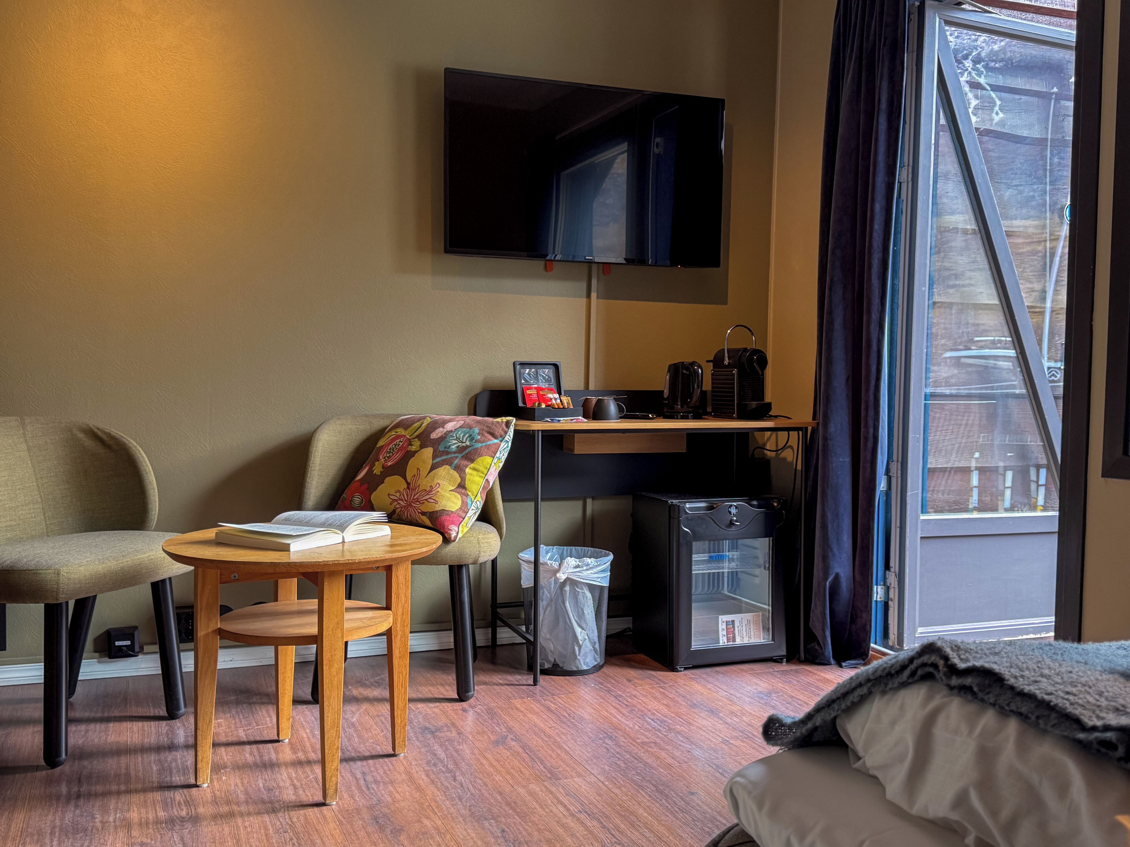 Et hotelværelse med en seng, et skrivebord med et fjernsyn, en stol, et lille bord med en bog, en skraldespand og et vindue med gardiner.