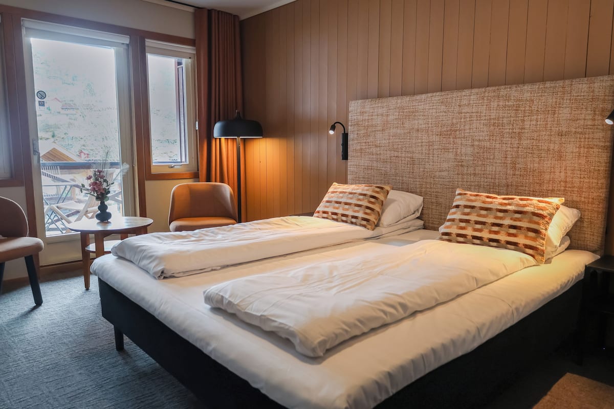 Una cama doble con ropa blanca, una mesita redonda con un florero y una silla.