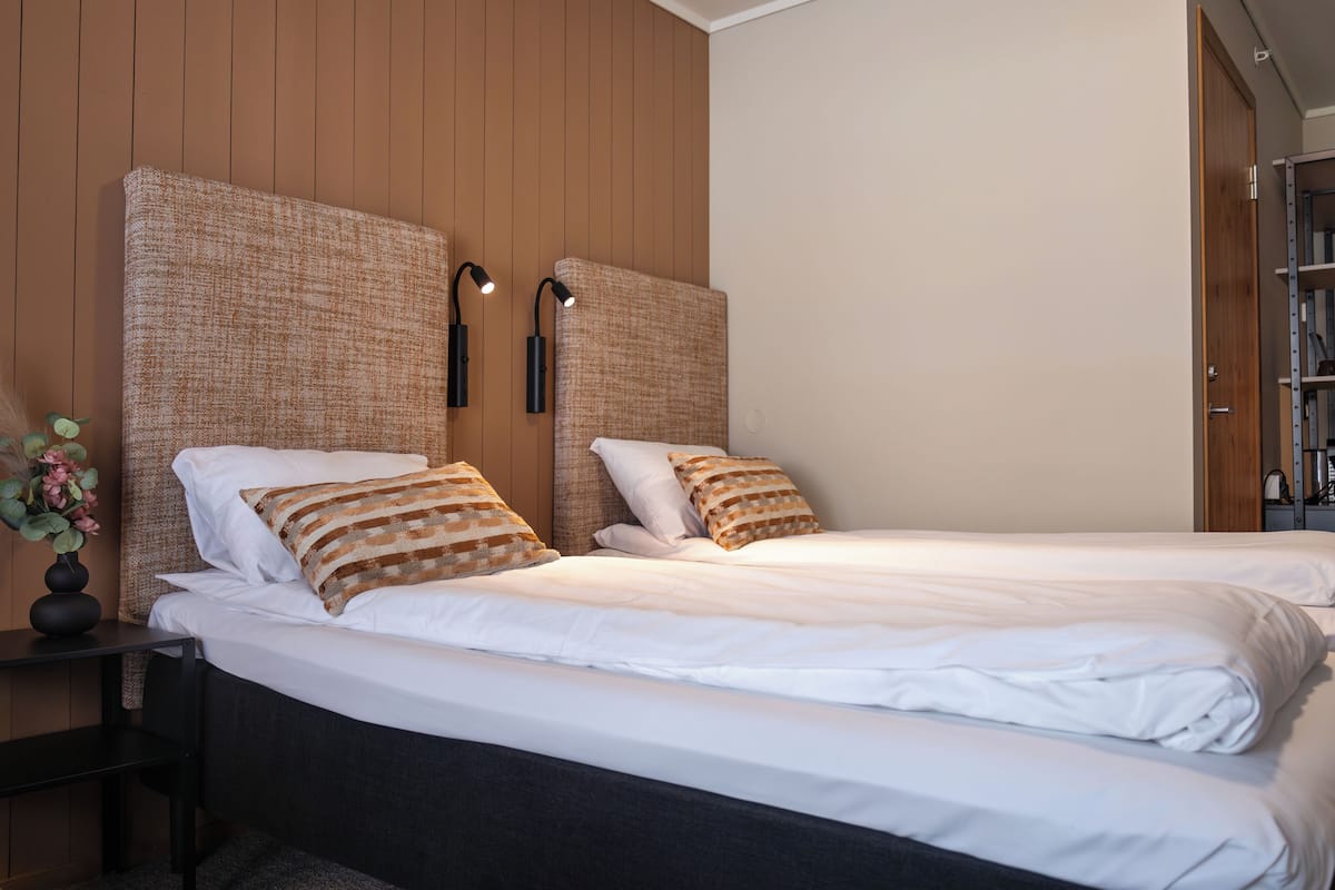 Una cama doble con sábanas blancas y almohadas estampadas, una mesita de noche con un jarrón de flores y un cabecero de madera con iluminación en la pared.