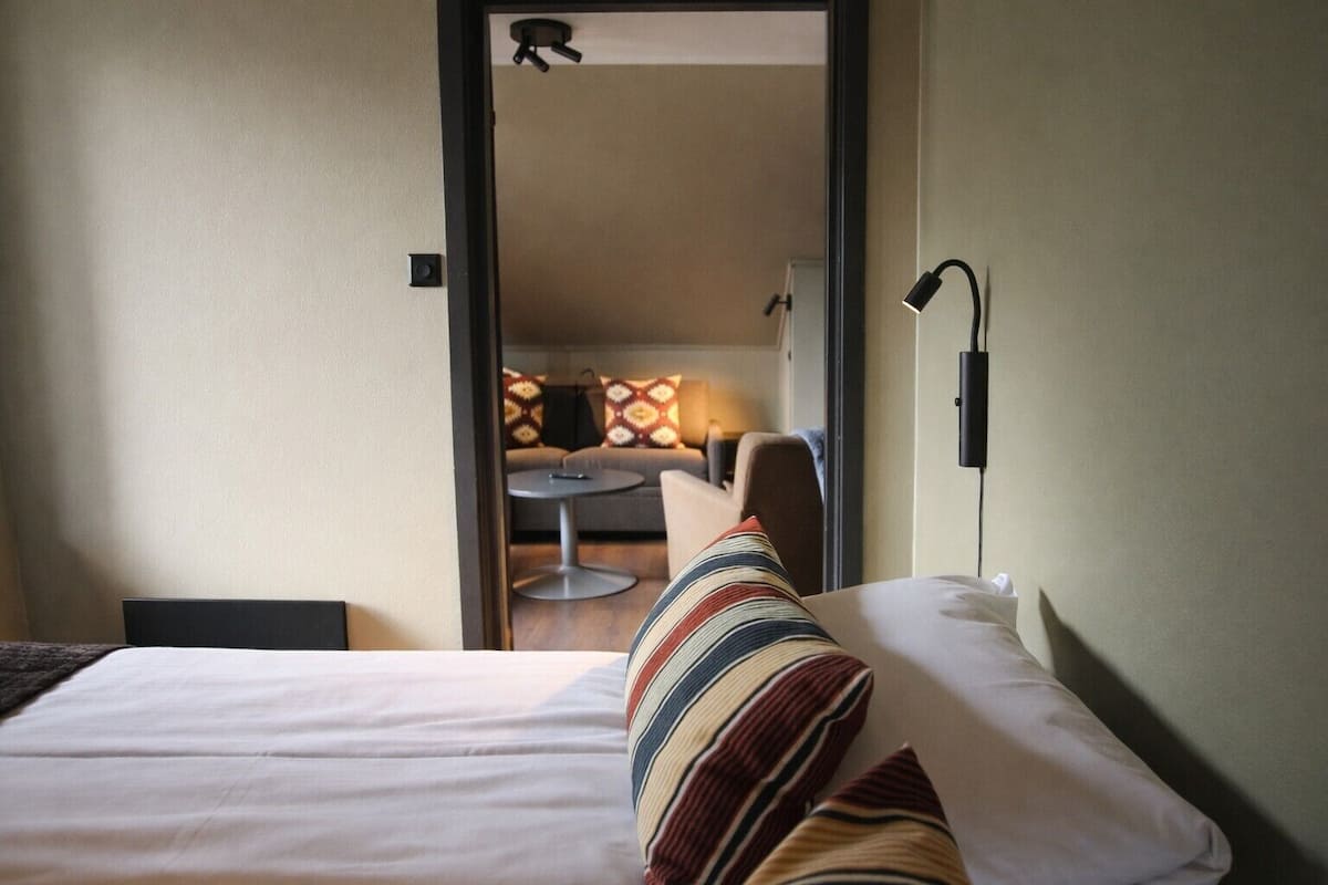 Habitación de hotel con una cama, una mesita de noche, una lámpara y vistas a un área de estar con sofá y mesa.