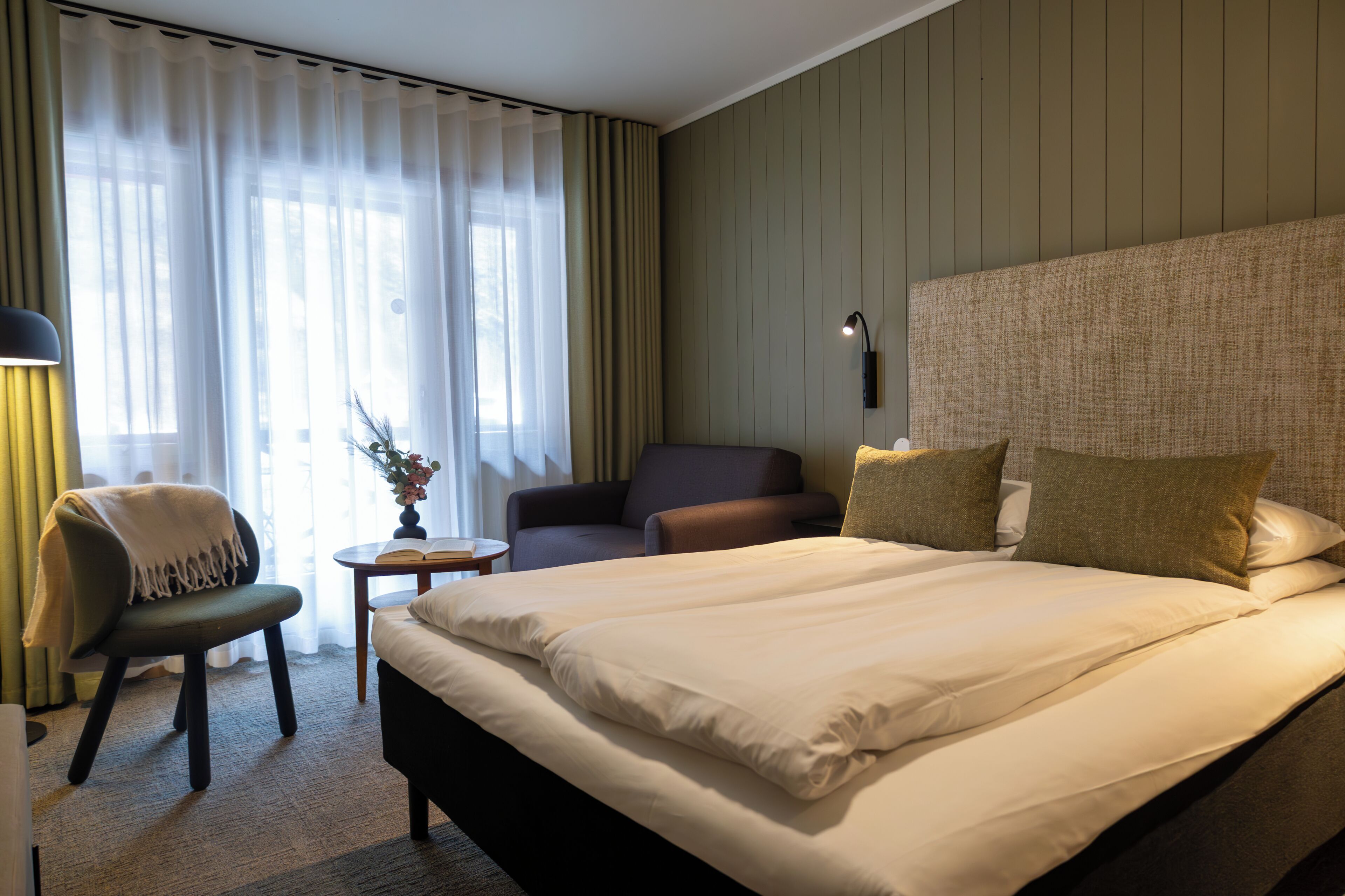 A hotel room with a large bed, a chair, a small table, and a lamp.
