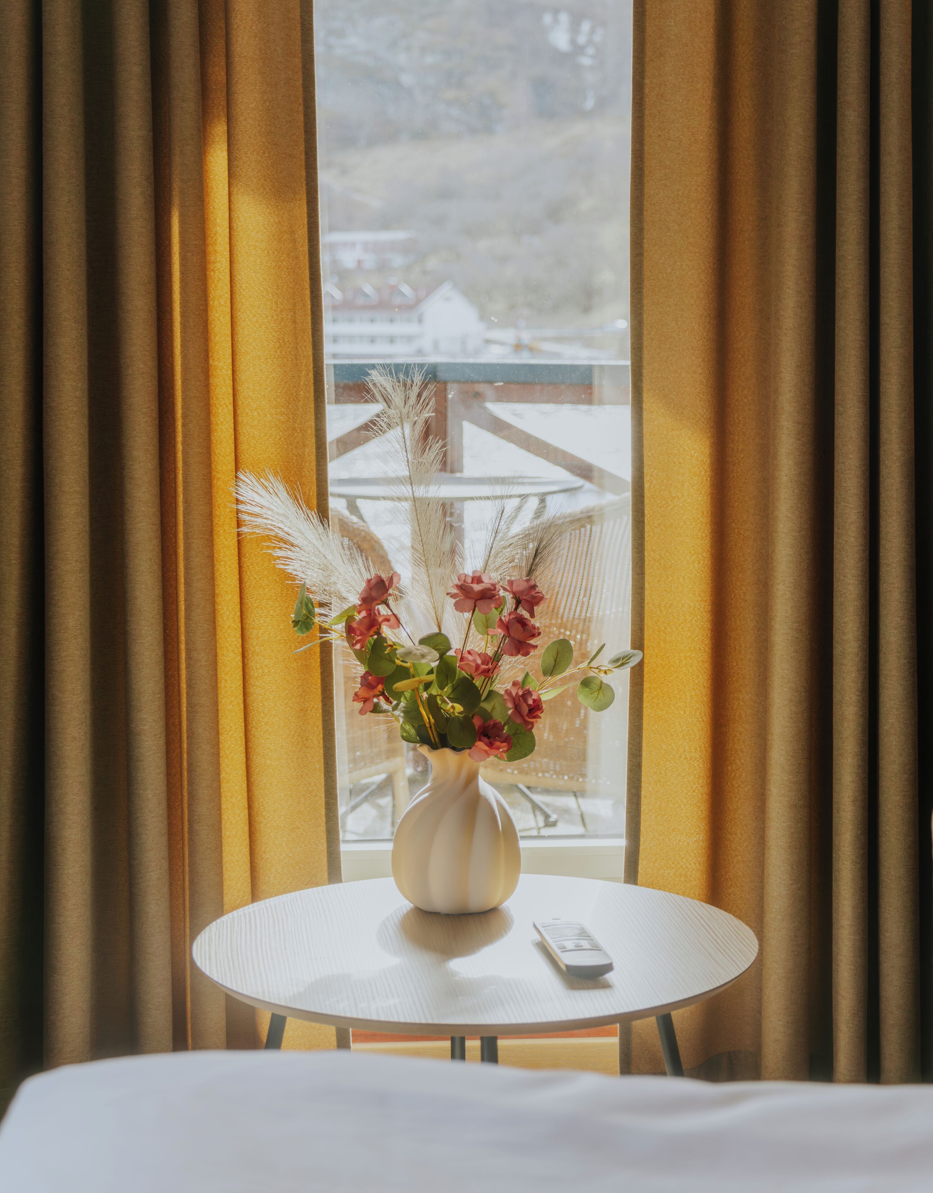 Una habitación con una mesa redonda, un jarrón con flores y vistas a través de una ventana con cortinas.