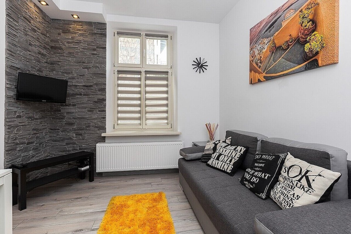 Uma sala de estar moderna com parede de pedra, uma janela grande com persianas, um sofá cinza com almofadas decorativas e um tapete amarelo.
