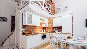 A kitchen with white cabinets, a brick wall, a wooden staircase, and a dining area with white chairs and a table. - EchoDom 3 Apartments (Kraków)