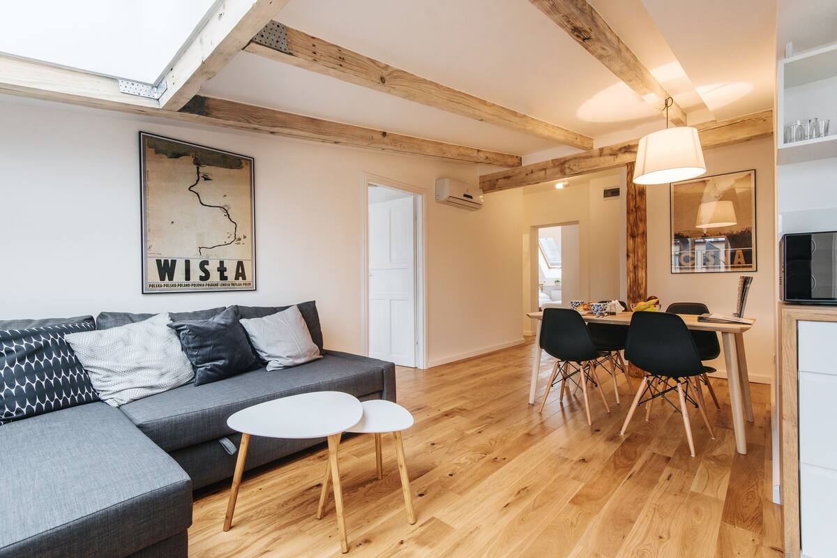 Uma sala de estar moderna com um sofá cinzento, duas mesas de apoio brancas, piso de madeira, uma área de jantar com mesa e cadeiras, e uma cozinha com luminária pendente.
