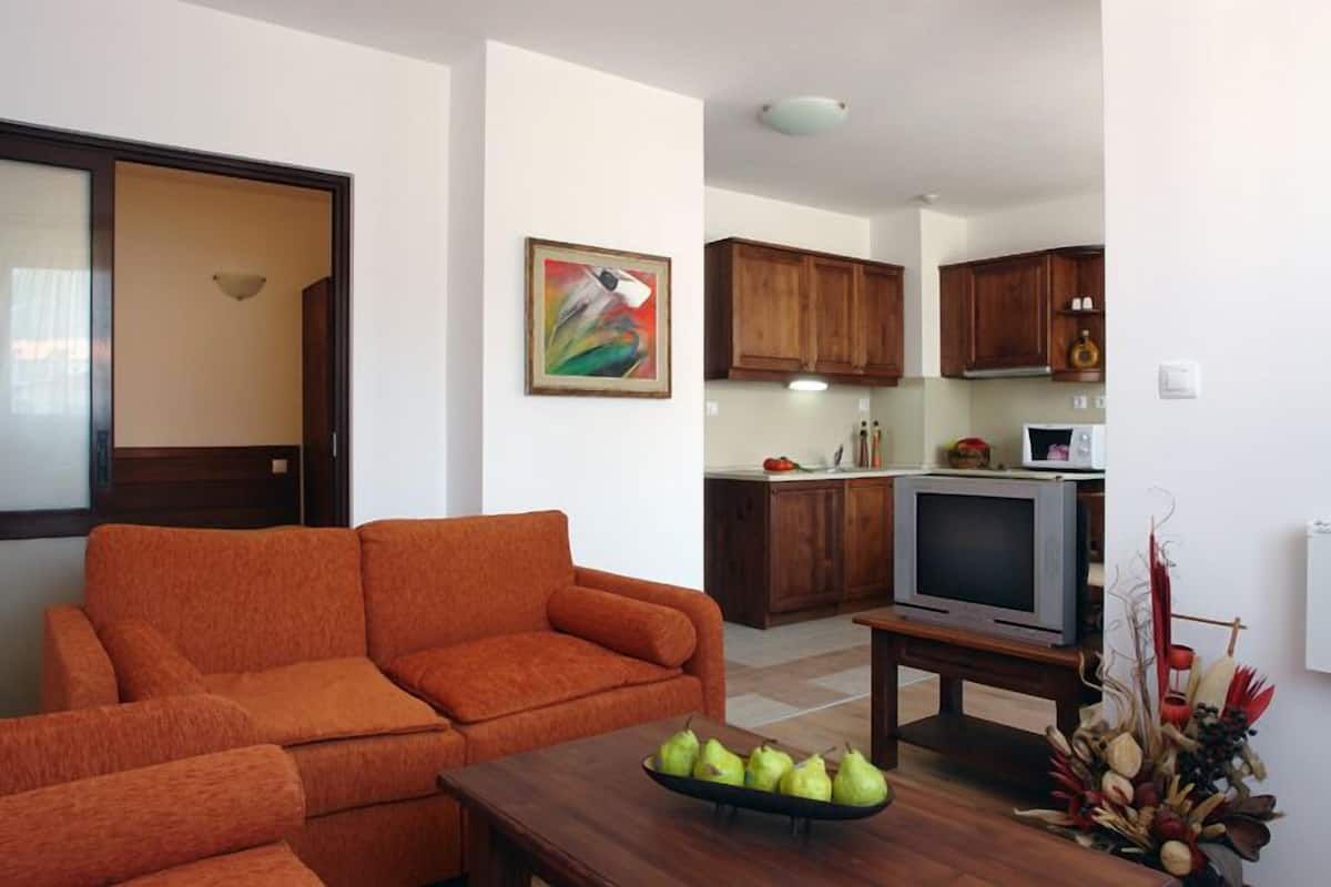 A living room with a sofa, a coffee table with pears, a television on a stand, and a kitchen in the background.