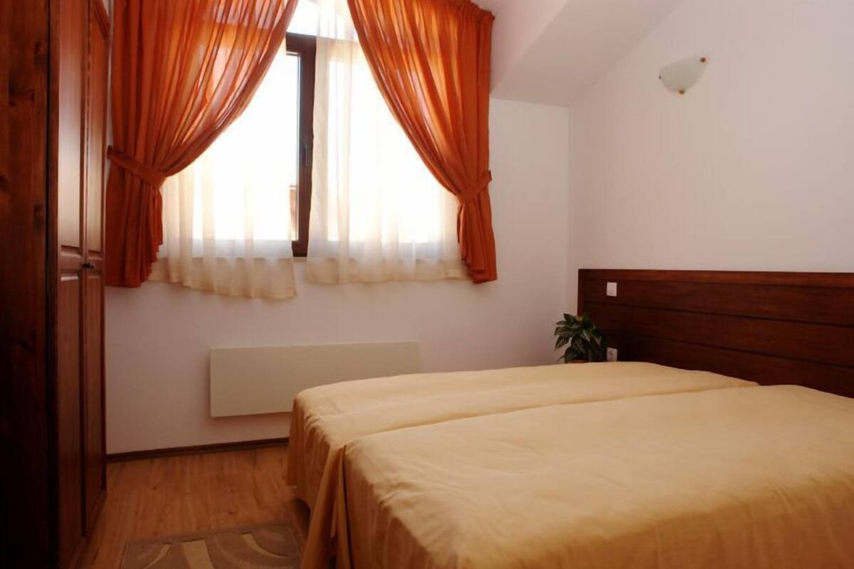 Un dormitorio con cama doble, ventana con cortinas traslúcidas, radiador y una planta en la mesita de noche.
