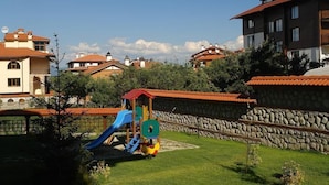 A playground with slides and climbing structures, surrounded by stone walls and residential buildings with red-tiled roofs. - Winslow Elegance Hotel (Bansko)