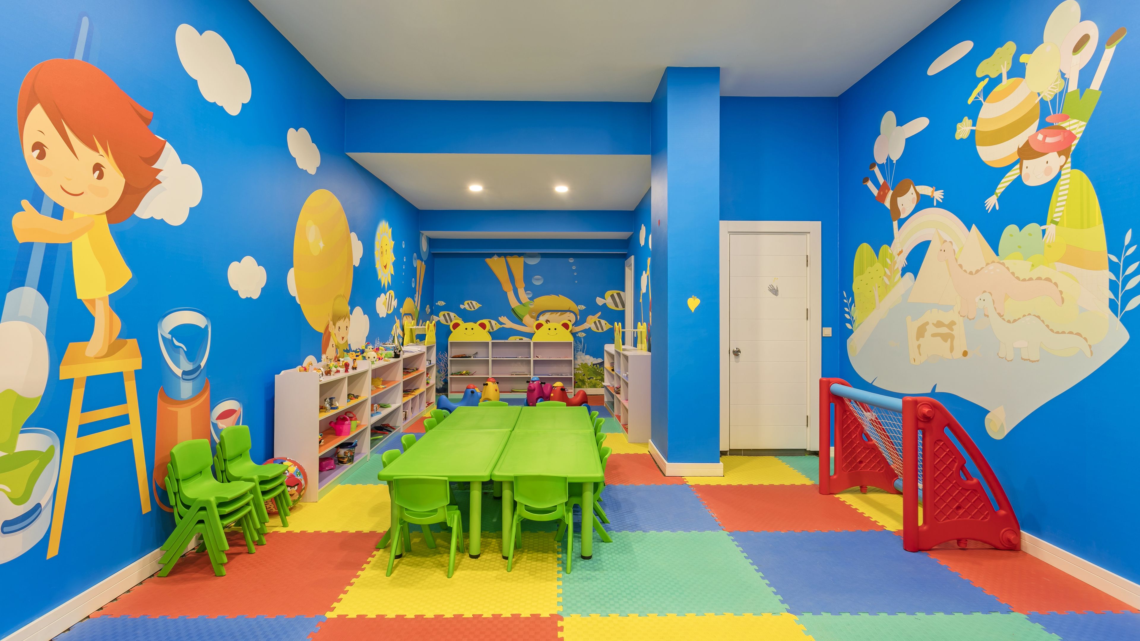 A children's playroom with a blue wall, colorful floor mats, green tables, and shelves filled with toys.