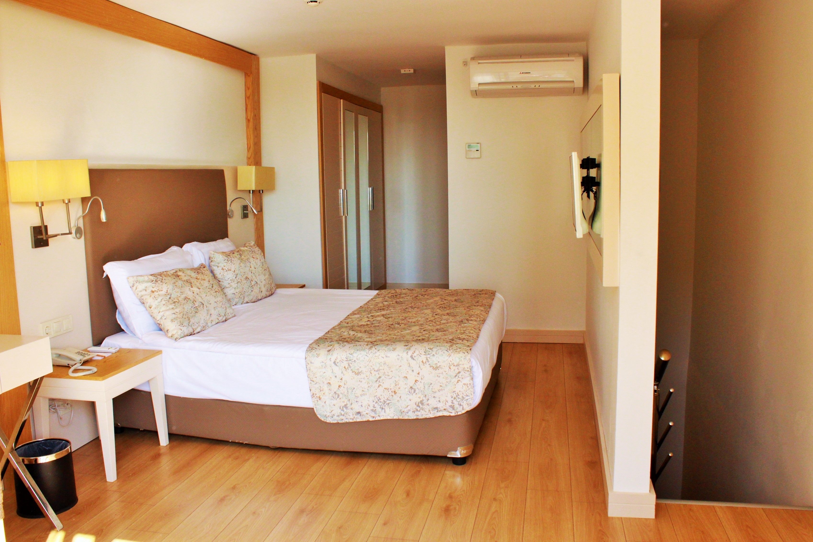 A hotel room with a bed, bedside table, lamp, and an air conditioning unit.