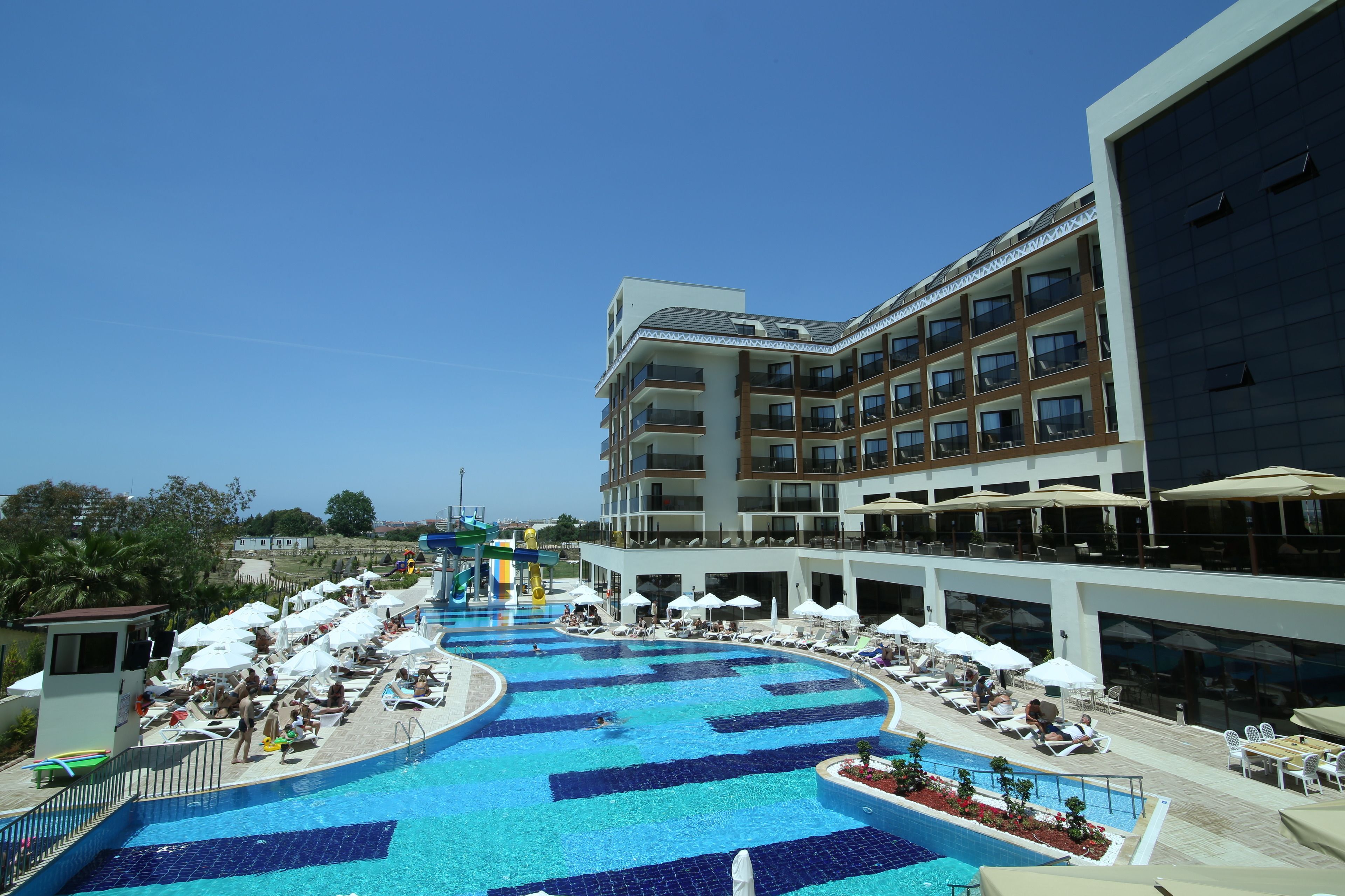 A large outdoor swimming pool with multiple lounge areas and umbrellas, surrounded by a modern hotel building with balconies and large windows.