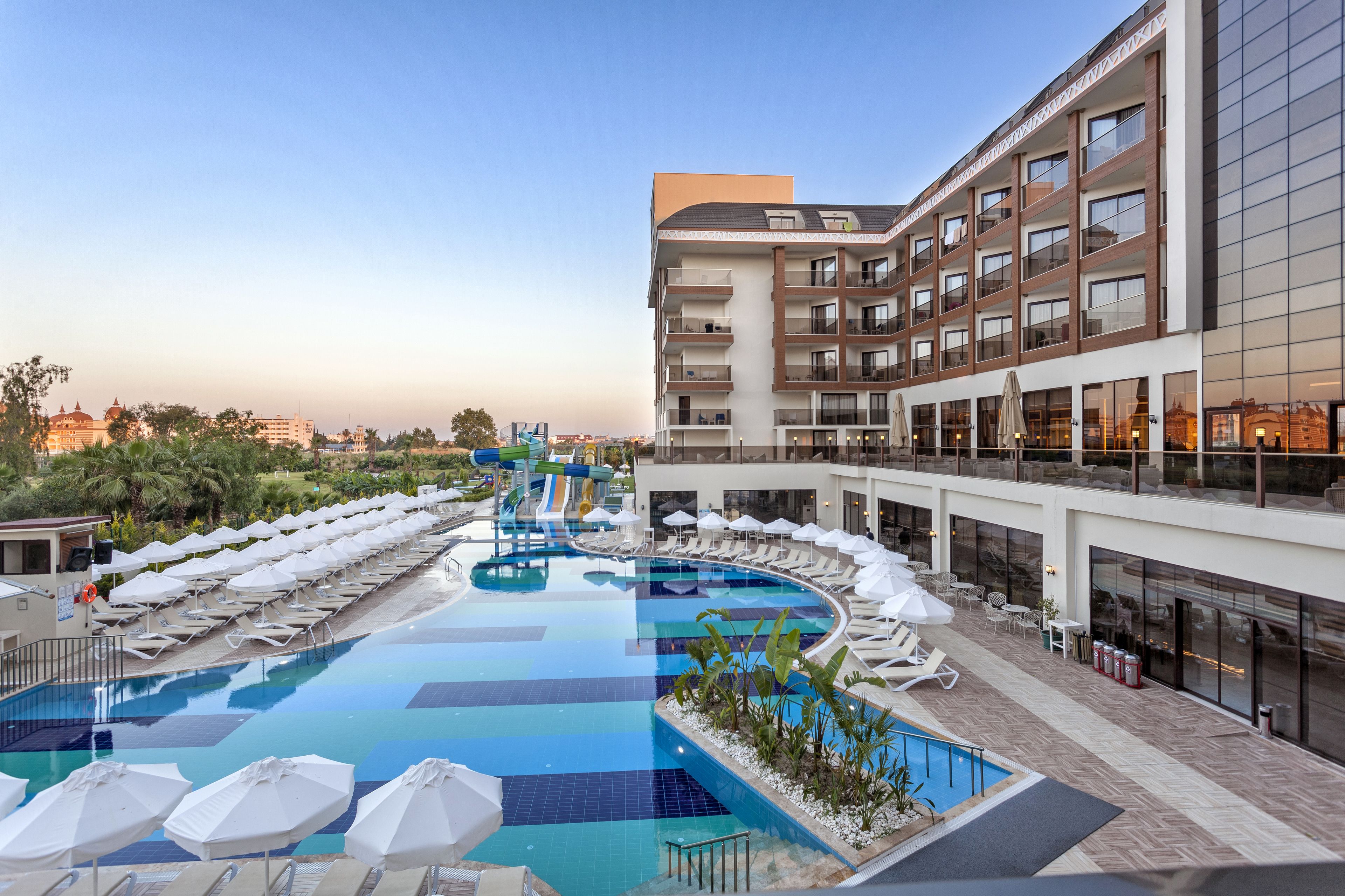 A rooftop pool with lounge chairs and umbrellas, a water slide, and a modern building with balconies.