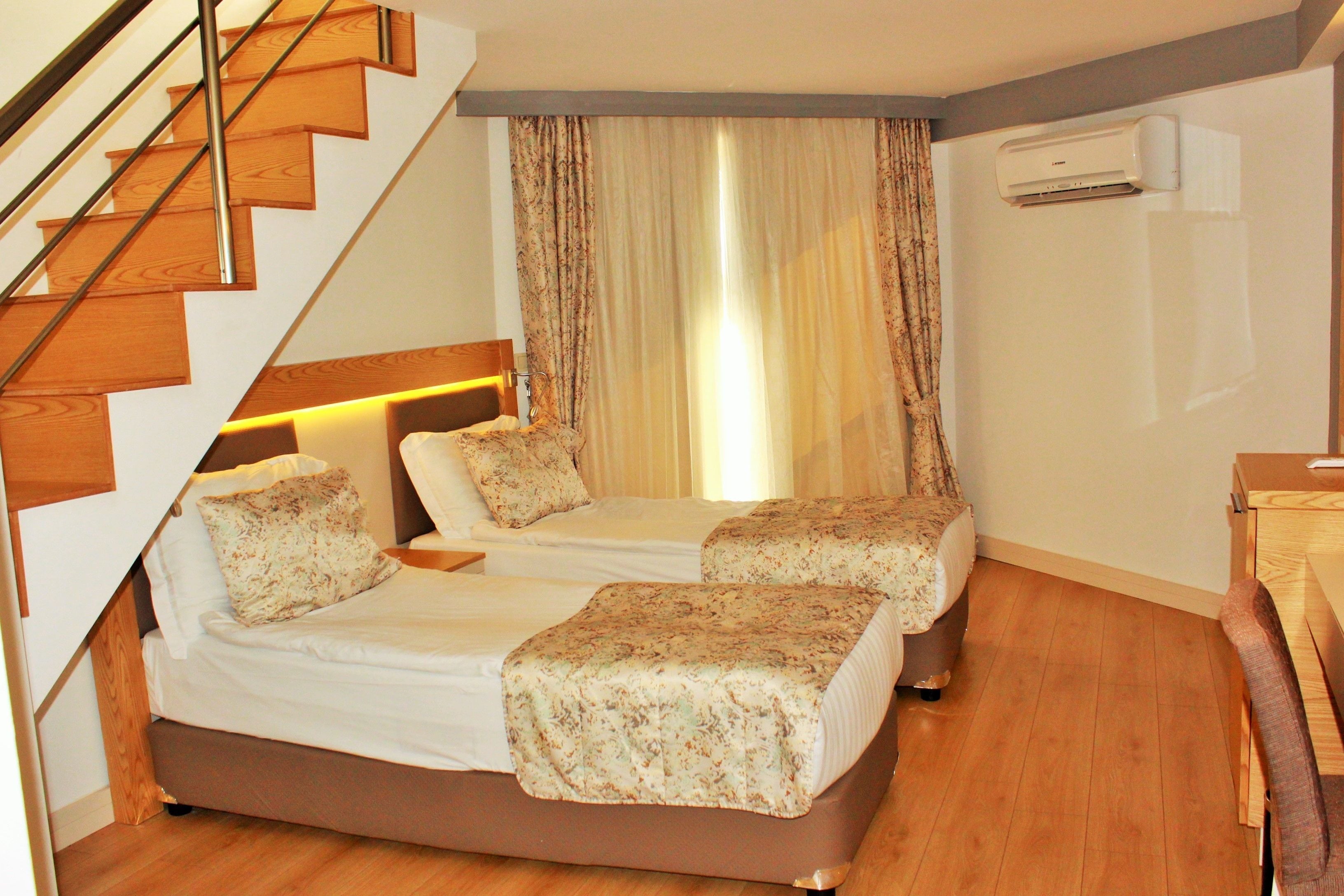 A bedroom with a wooden staircase, two beds with floral bedspreads, a bedside table, a chair, and an air conditioning unit.