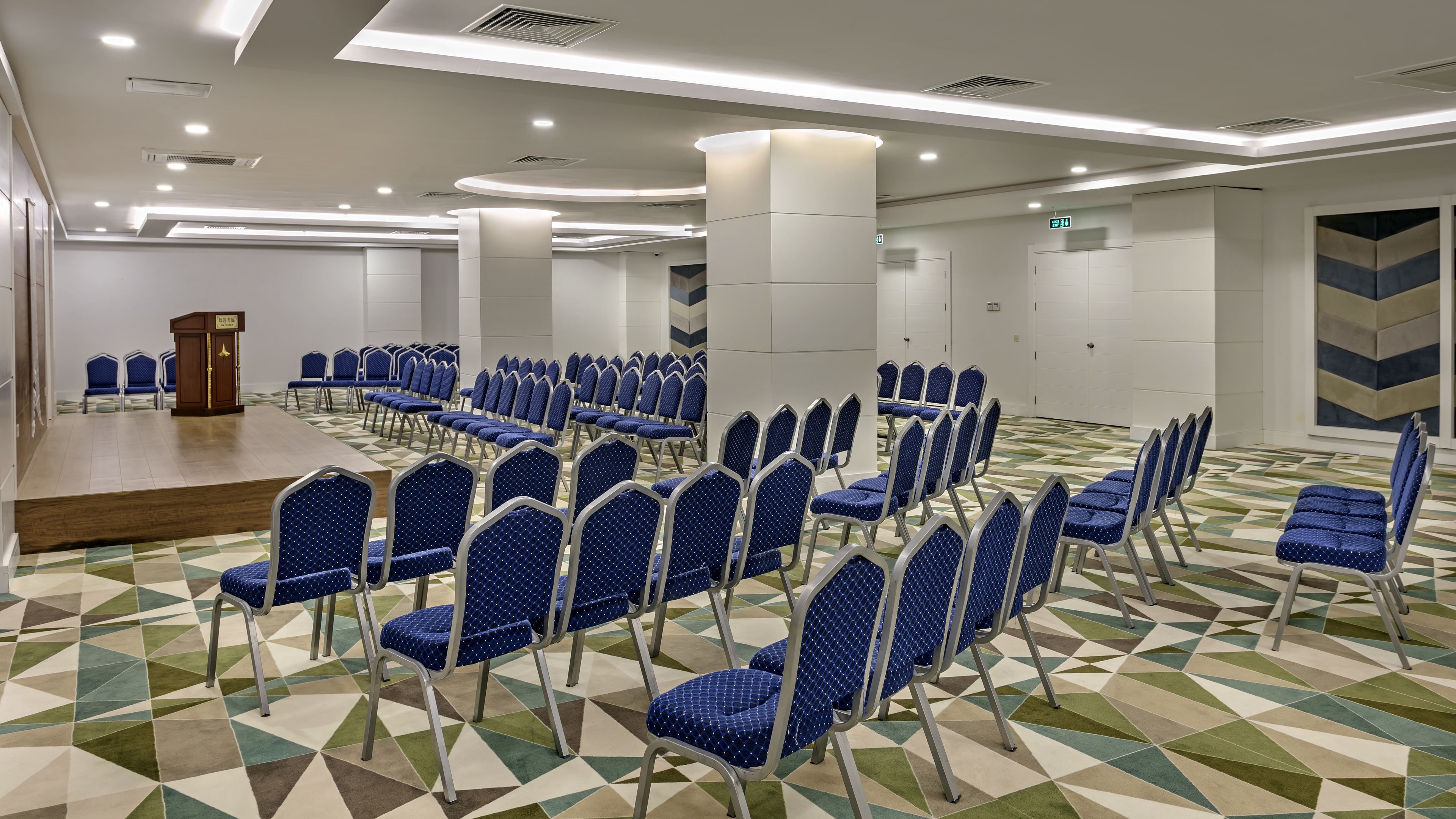 A conference room with rows of blue chairs, a stage, and a patterned carpet.