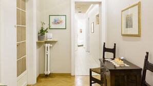A narrow hallway with a wooden floor, a radiator, and a dining table with chairs. There are framed pictures on the walls and a plant on a shelf. - Relais Calamatta (Rome)